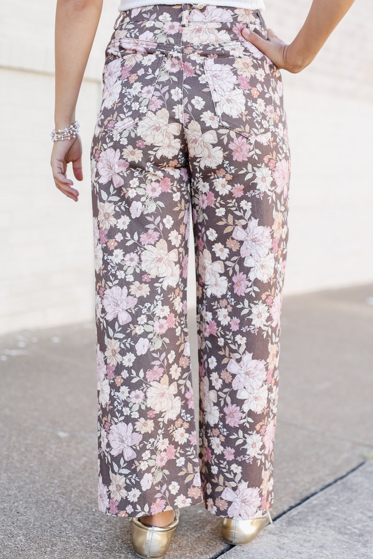 A person wearing high-waisted, wide-leg pants with a brown base and a pink, white, and beige floral pattern, paired with metallic gold shoes.