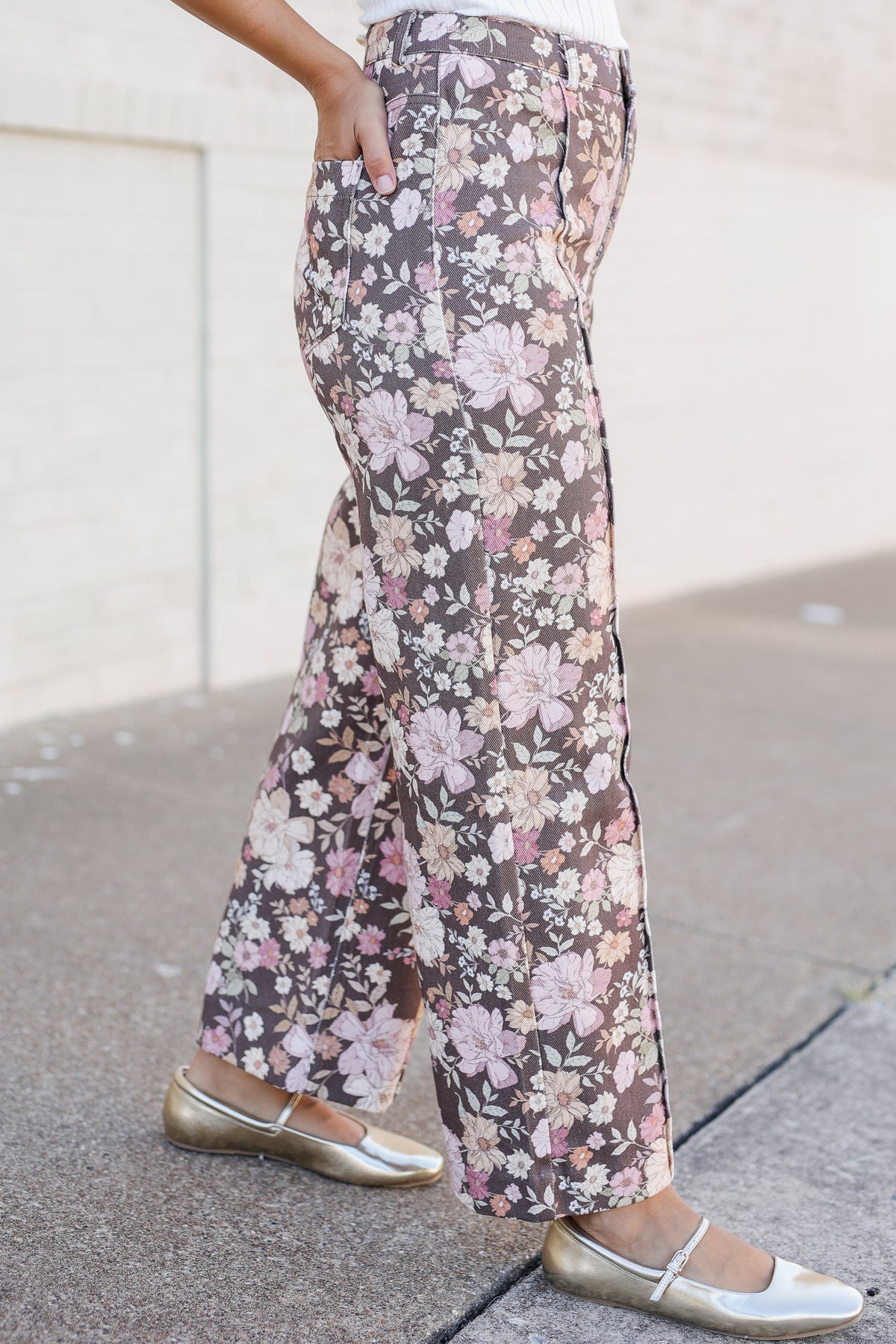A person wearing wide-leg floral pants with a brown base and pink, white, and beige flowers, paired with metallic gold flats, standing on a sidewalk.