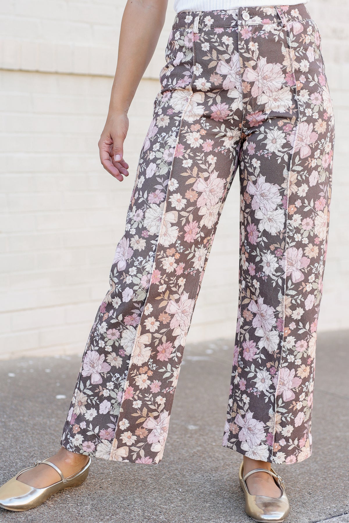 A person wearing high-waisted, wide-leg floral pants with a brown base and pink, white, and green floral pattern, paired with metallic gold flats, standing against a light brick wall.