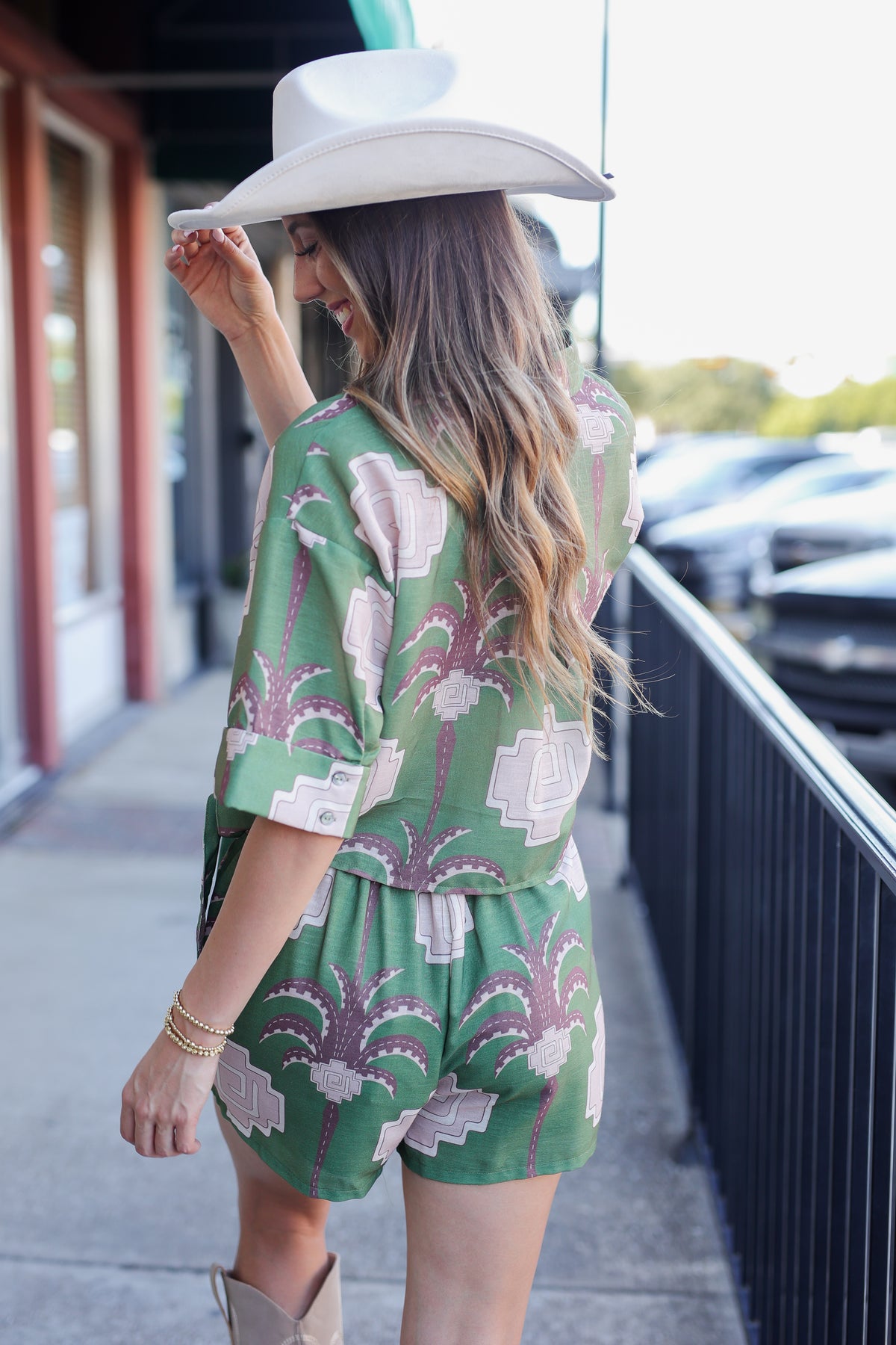 Woman wearing a green patterned romper and white cowboy hat on a city street.