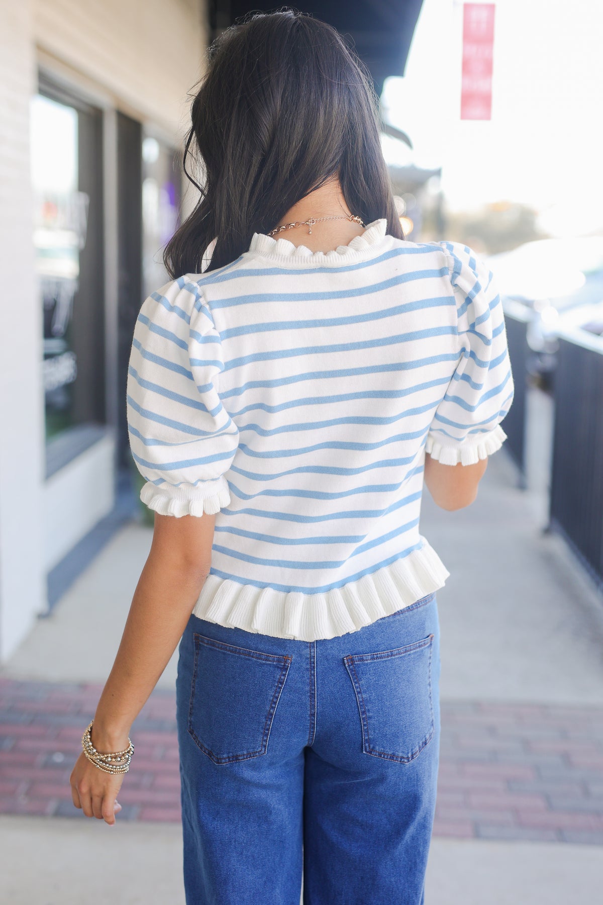 Person wearing a blue and white striped shirt with ruffled details and blue jeans, standing on a sidewalk.