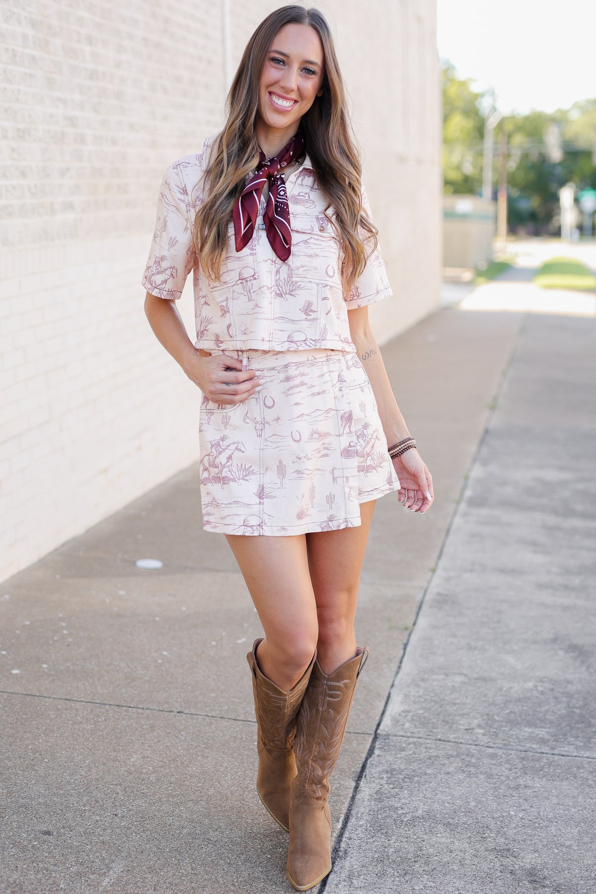 A woman stands on a sidewalk wearing a matching short-sleeve top and mini skirt set in a light pink and white botanical print, styled with a dark red neck scarf and brown cowboy boots.