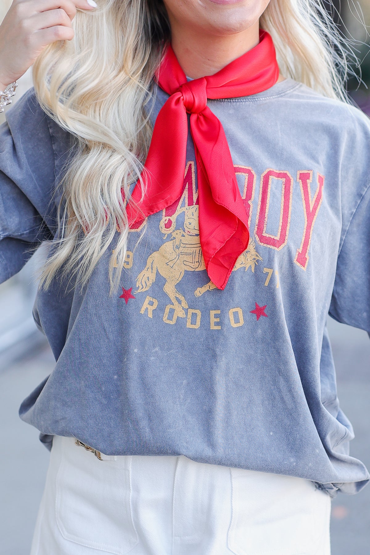 A close-up of a person wearing a faded slate blue graphic t-shirt with a red neck scarf and white pants. The shirt features a distressed gold and red 'COWBOY RODEO' graphic.