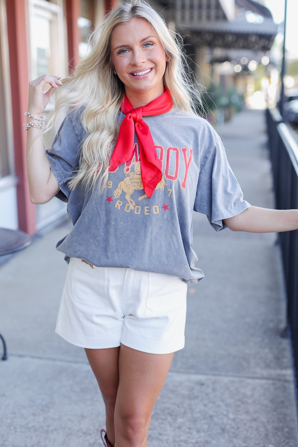 A smiling blonde woman stands outdoors wearing a faded gray graphic T-shirt with red lettering and a red neck scarf, paired with white shorts.