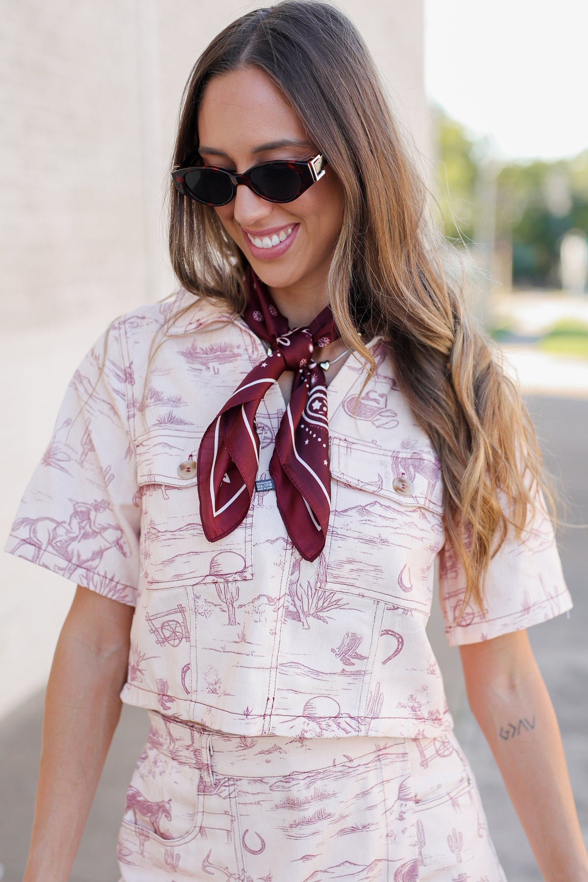 A woman wearing a matching short-sleeve button-up shirt and shorts set in a cream color with a dusty rose toile pattern, accessorized with a dark red paisley neck scarf and black sunglasses.