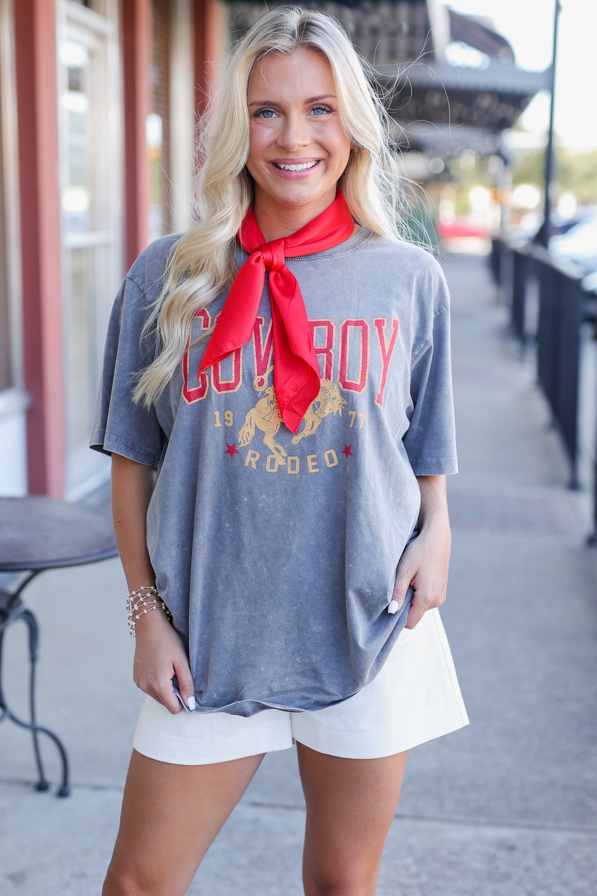 A smiling blonde woman stands outdoors wearing an oversized heather gray graphic T-shirt with red and gold 'Cowboy Rodeo' lettering, a bright red neck scarf, and white shorts.