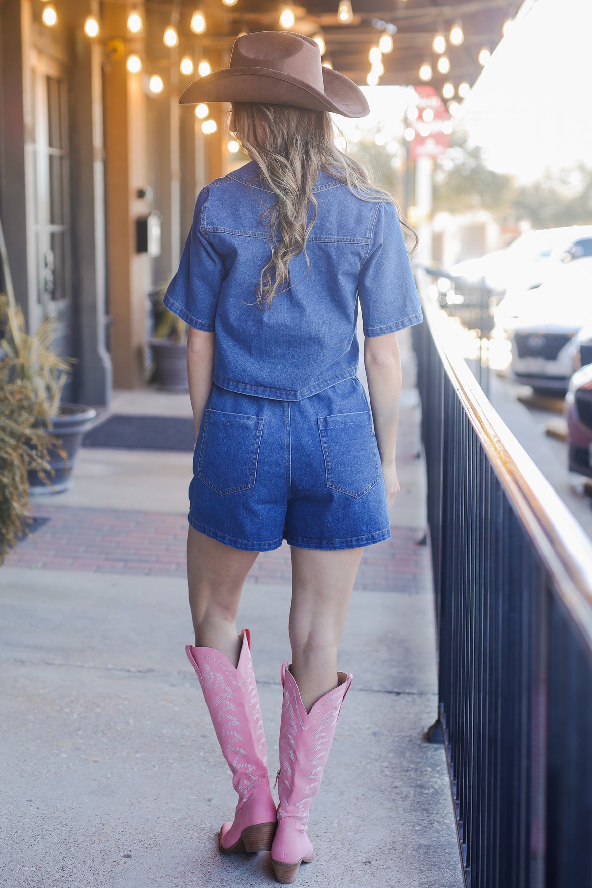 Woman wearing a blue denim romper and pink cowboy boots standing on a sidewalk.