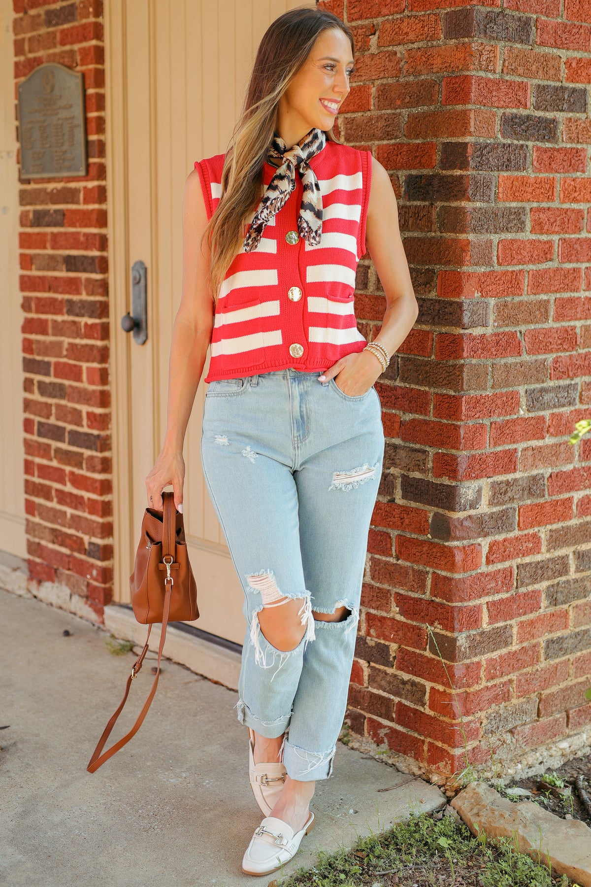 A woman stands outdoors against a brick wall and beige door, wearing a sleeveless red-and-white striped top with a black-and-white patterned scarf, light-wash ripped jeans, white flats, and carrying a brown handbag.