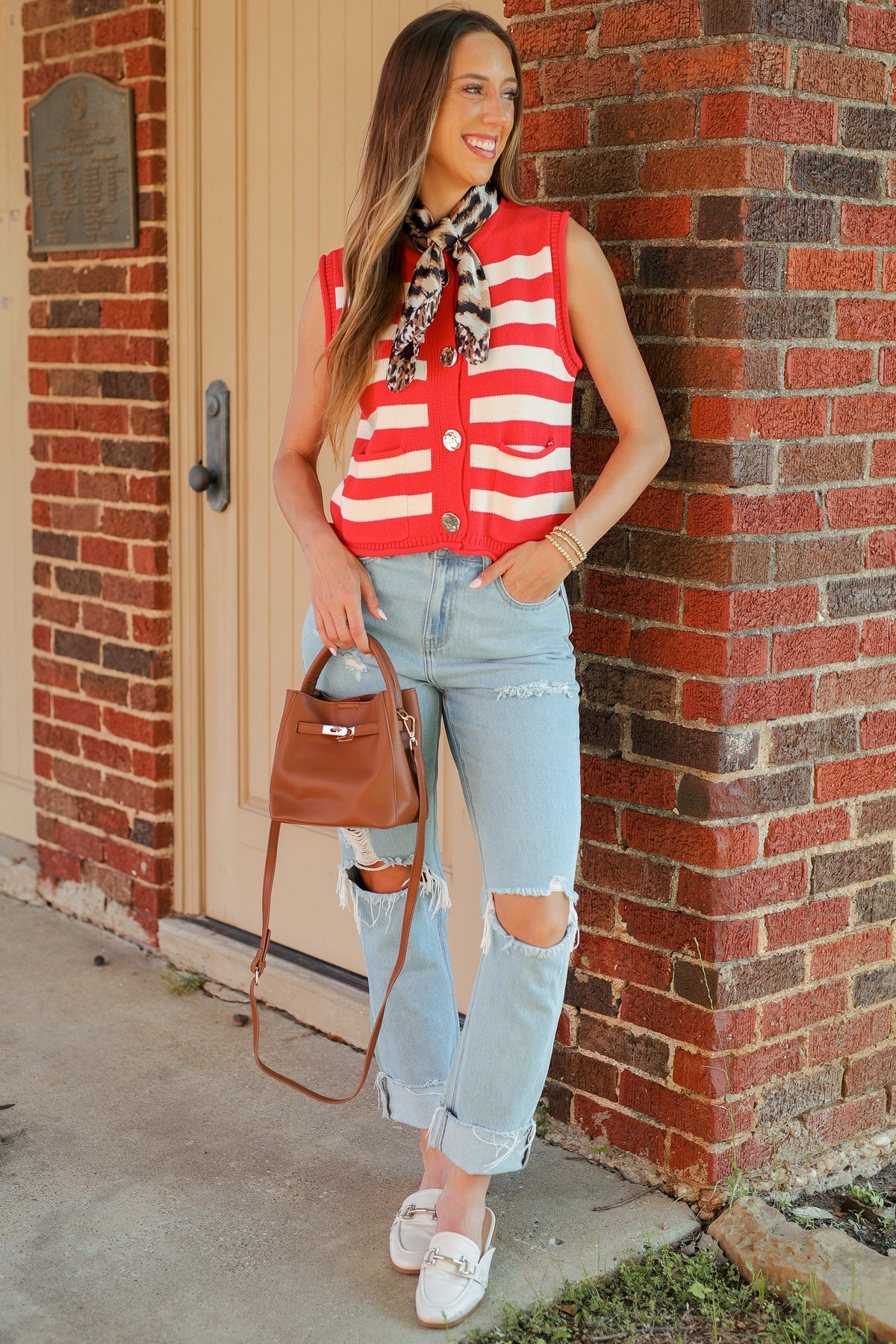A woman stands outdoors against a red brick wall wearing a red and white striped sleeveless top, light-wash ripped jeans, white loafers, and a brown handbag.