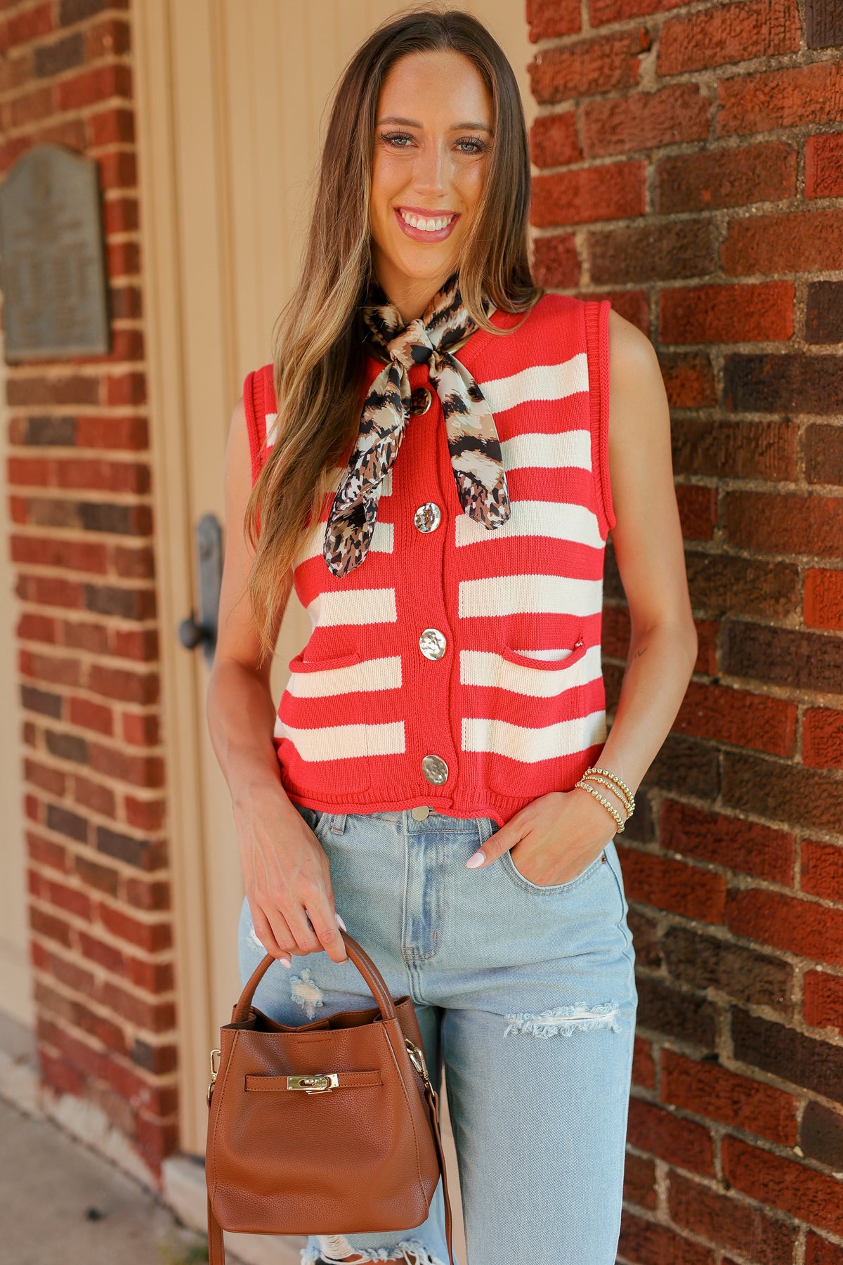 A woman stands against a red brick wall wearing a sleeveless red and white striped vest with a leopard-print neck scarf, light-wash distressed jeans, and a brown leather handbag.