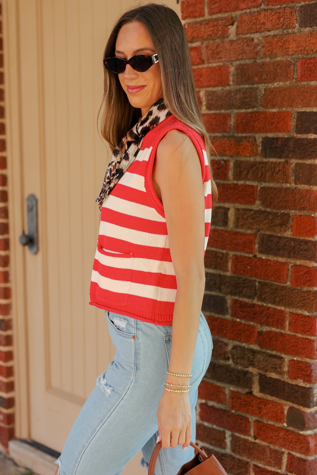 A woman wearing a red and white striped sleeveless knit top, light blue jeans, and a leopard print scarf stands in front of a brick wall.