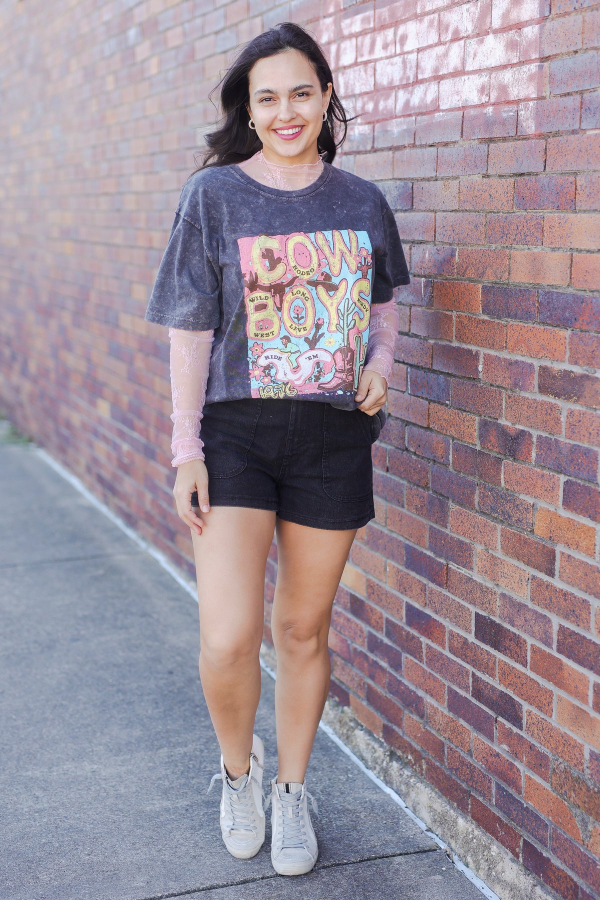 Woman wearing a graphic t-shirt and shorts standing against a brick wall.