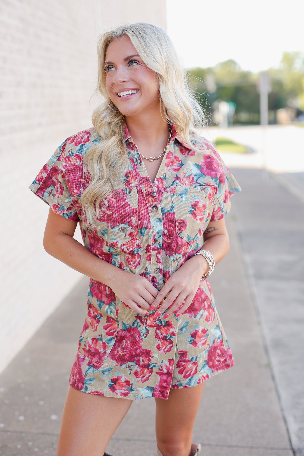 A woman with blonde hair wearing a short-sleeve floral romper, standing outdoors on a sidewalk next to a light-colored brick wall.