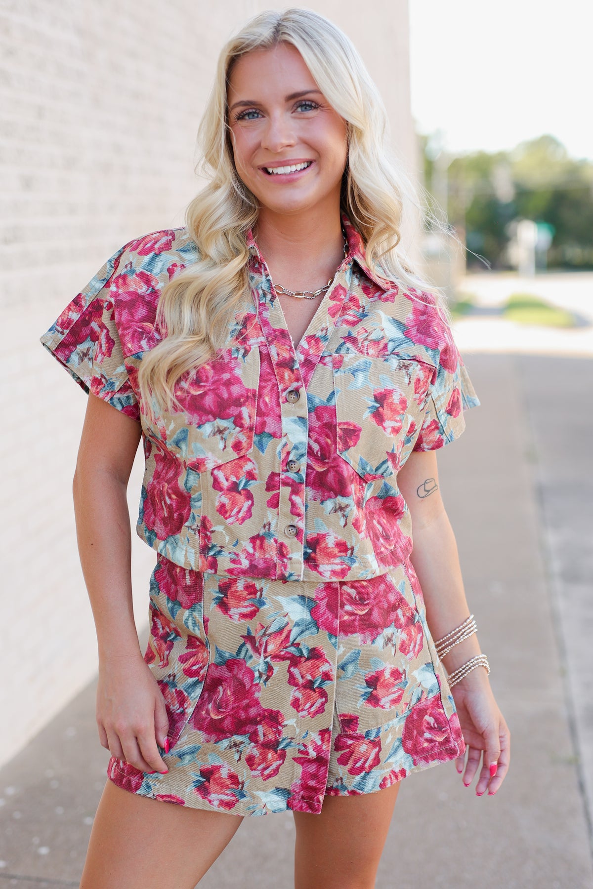 A woman modeling a matching short-sleeve button-front top and shorts set with a vibrant pink and red floral print on a beige background, standing outdoors against a light brick wall.
