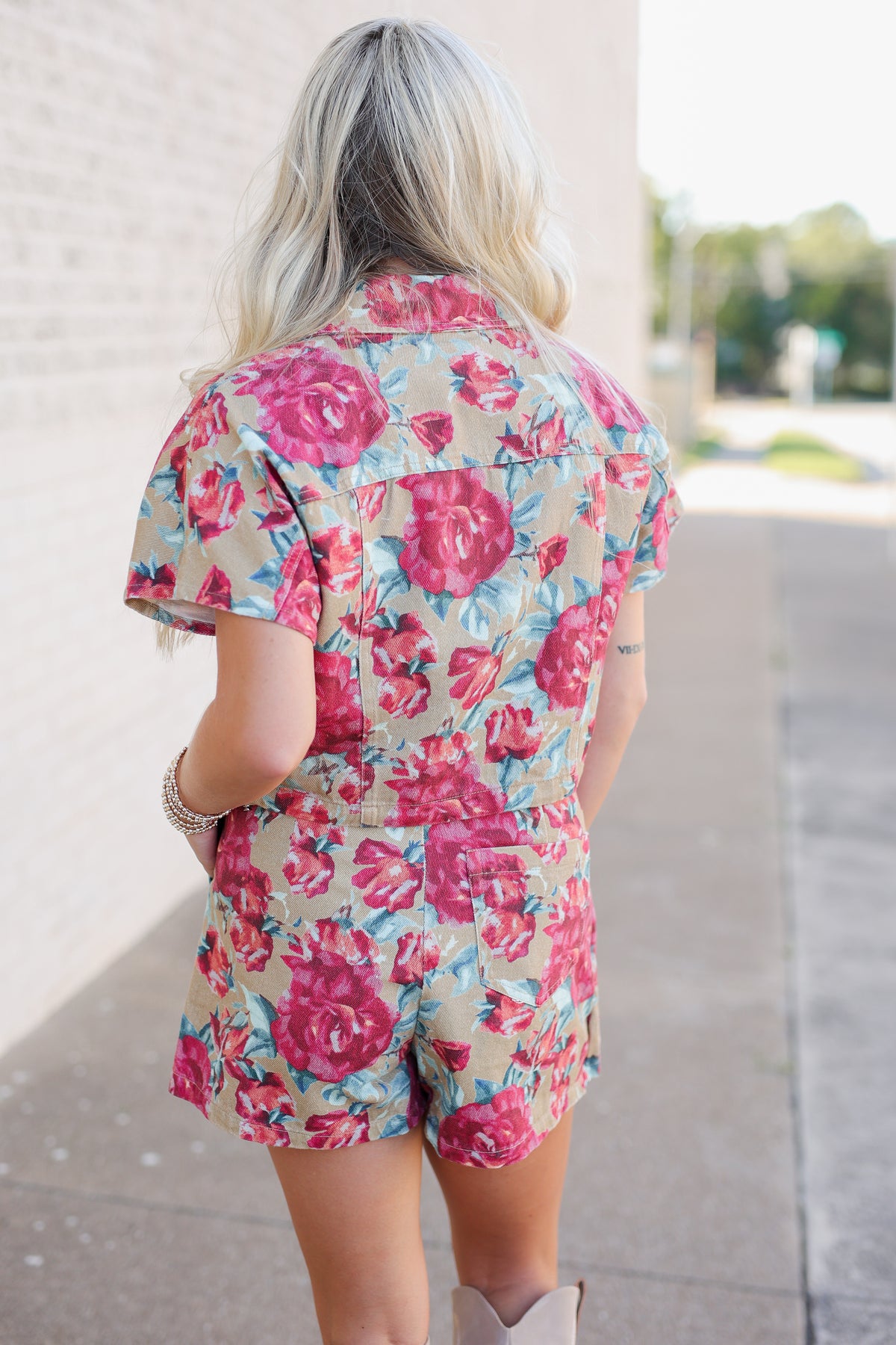 A woman viewed from behind wearing a short-sleeve floral romper with a beige base and large pink and red rose print, standing outdoors on a sidewalk next to a light brick wall.