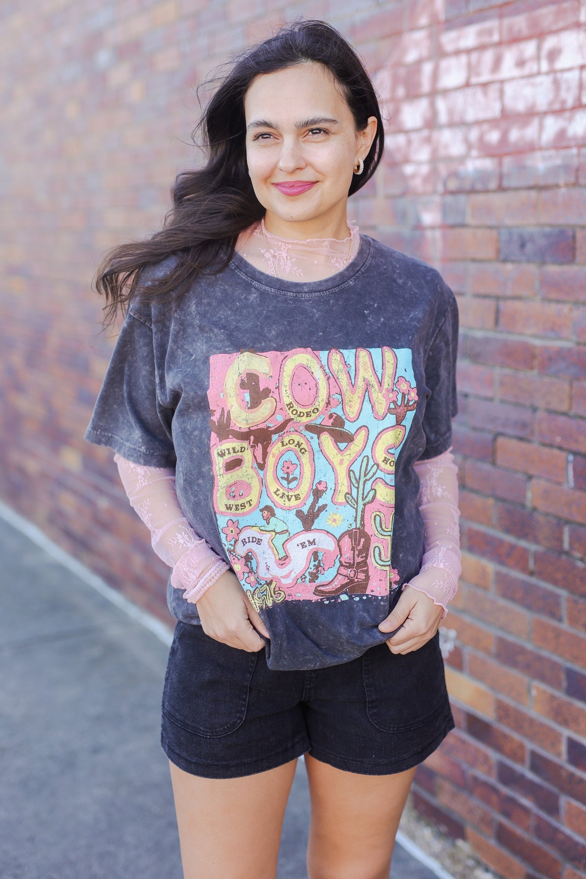 Woman wearing a graphic t-shirt with 'Cowboys' design against a brick wall.