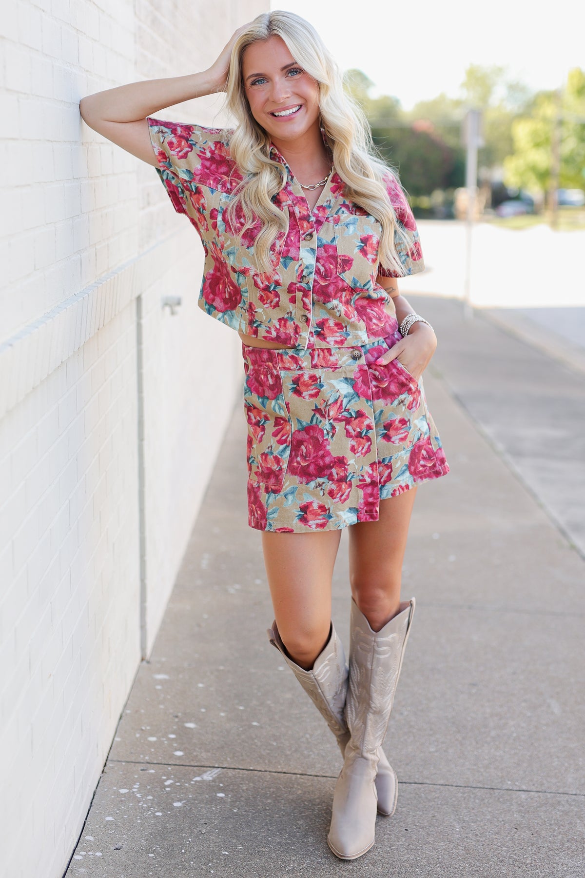 A woman stands on a sidewalk next to a white brick wall, wearing a short-sleeve button-front floral dress with a matching belt and light beige knee-high boots.