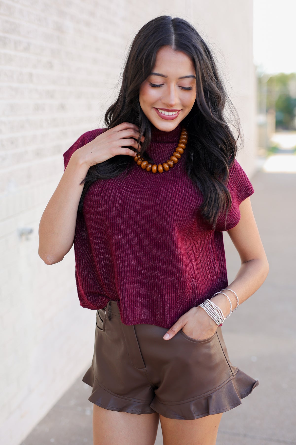 A woman wearing a short-sleeve ribbed knit top in deep burgundy and brown ruffled shorts, accessorized with a wooden bead necklace and silver bracelets, standing outdoors against a light brick wall.