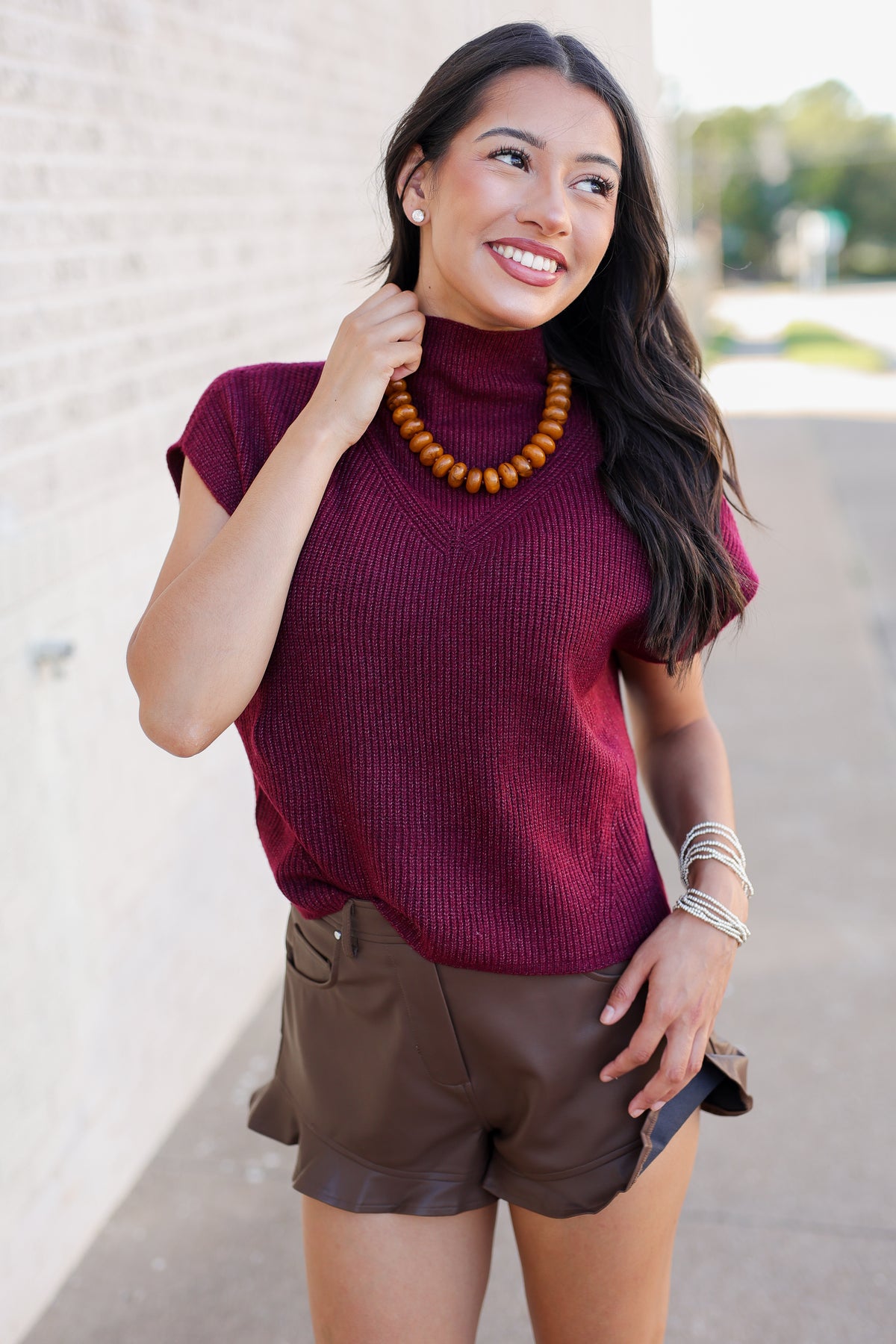A woman wearing a ribbed short-sleeve turtleneck sweater in deep burgundy and brown faux-leather shorts, accessorized with a wooden bead necklace and silver bracelets, standing outdoors against a light brick wall.