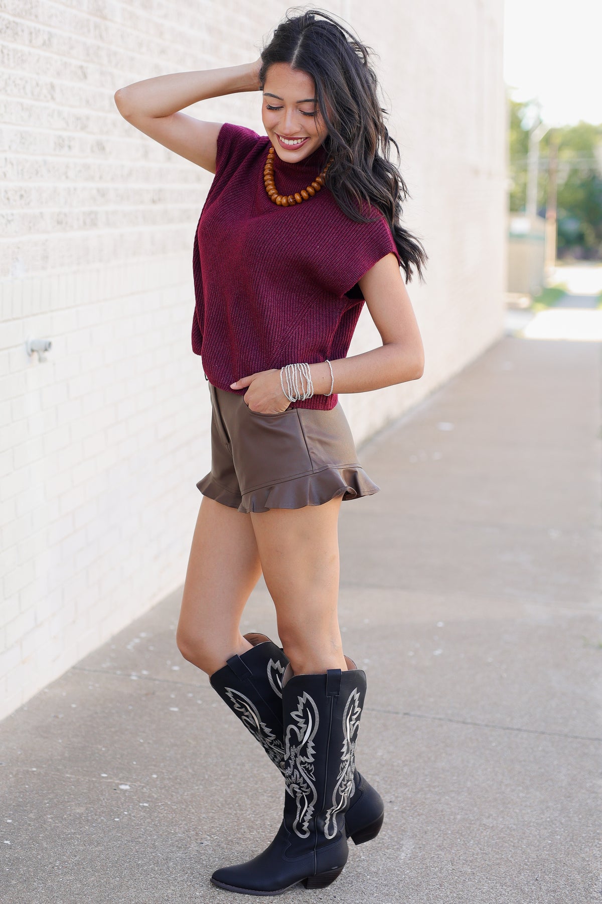 A woman posing outdoors in a maroon knit short-sleeve top, brown ruffled mini skirt, and black western boots with white embroidery.