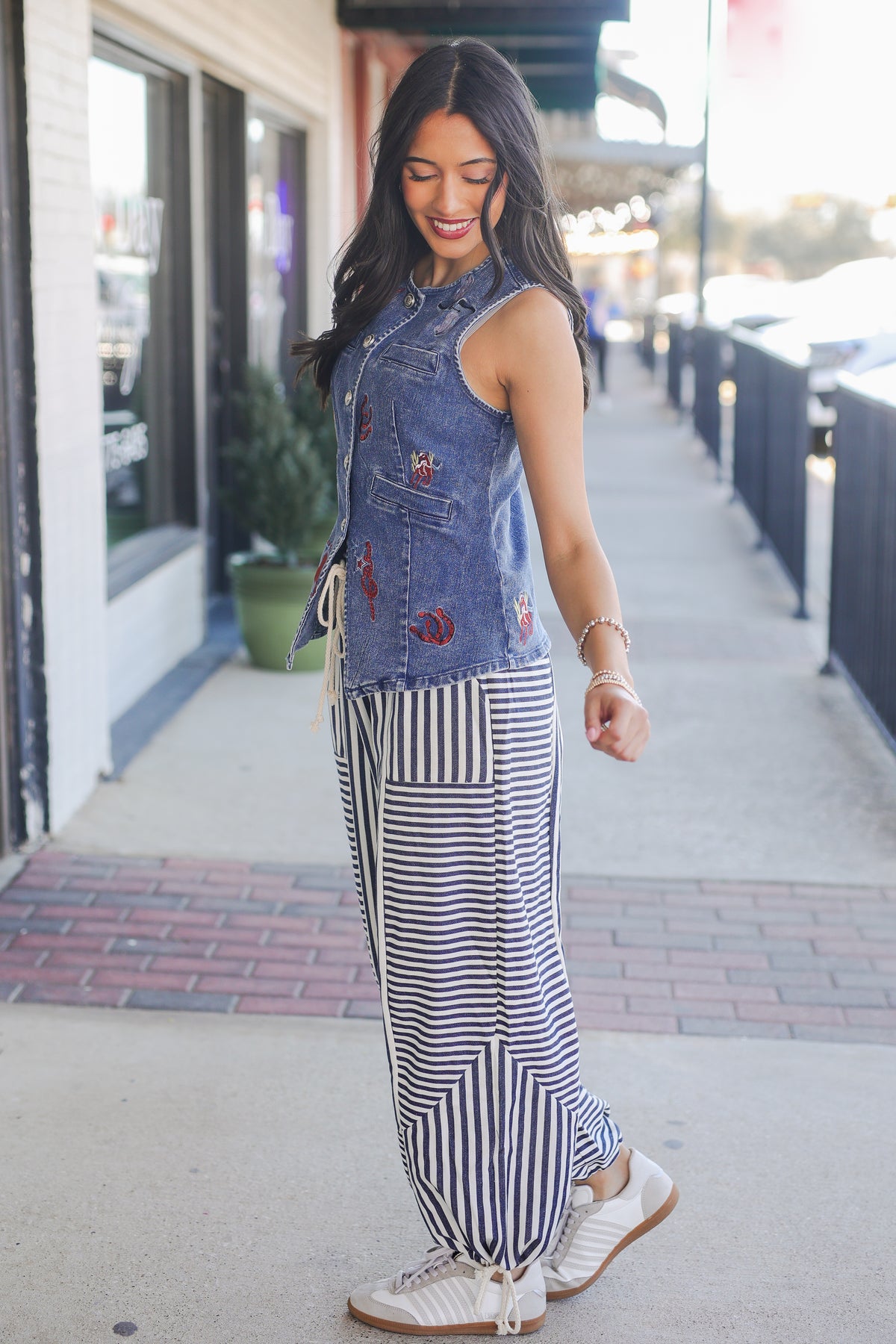 Woman wearing a denim vest with floral embroidery and striped pants on a sidewalk.