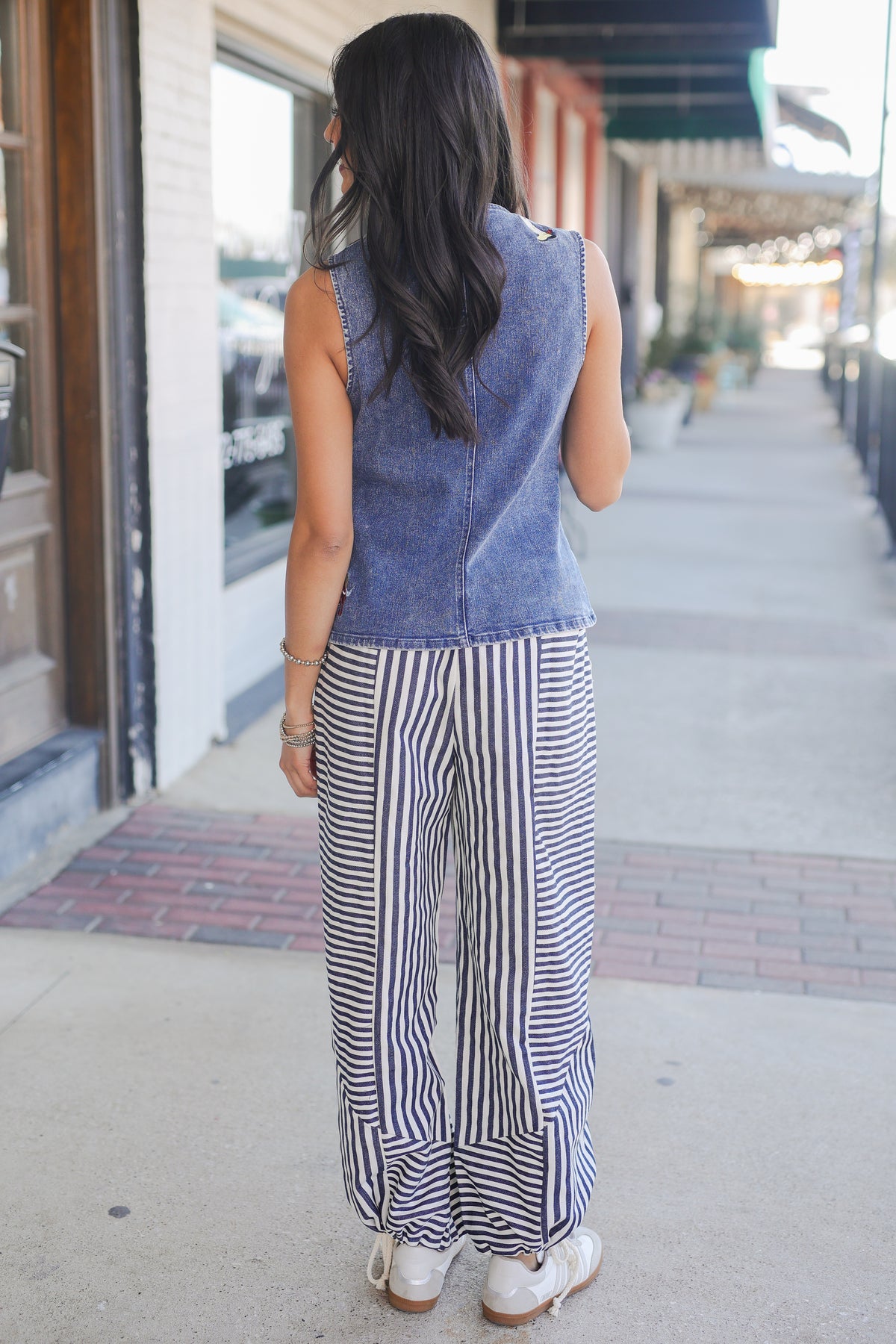 Woman wearing a denim sleeveless top and striped pants walking on a sidewalk.