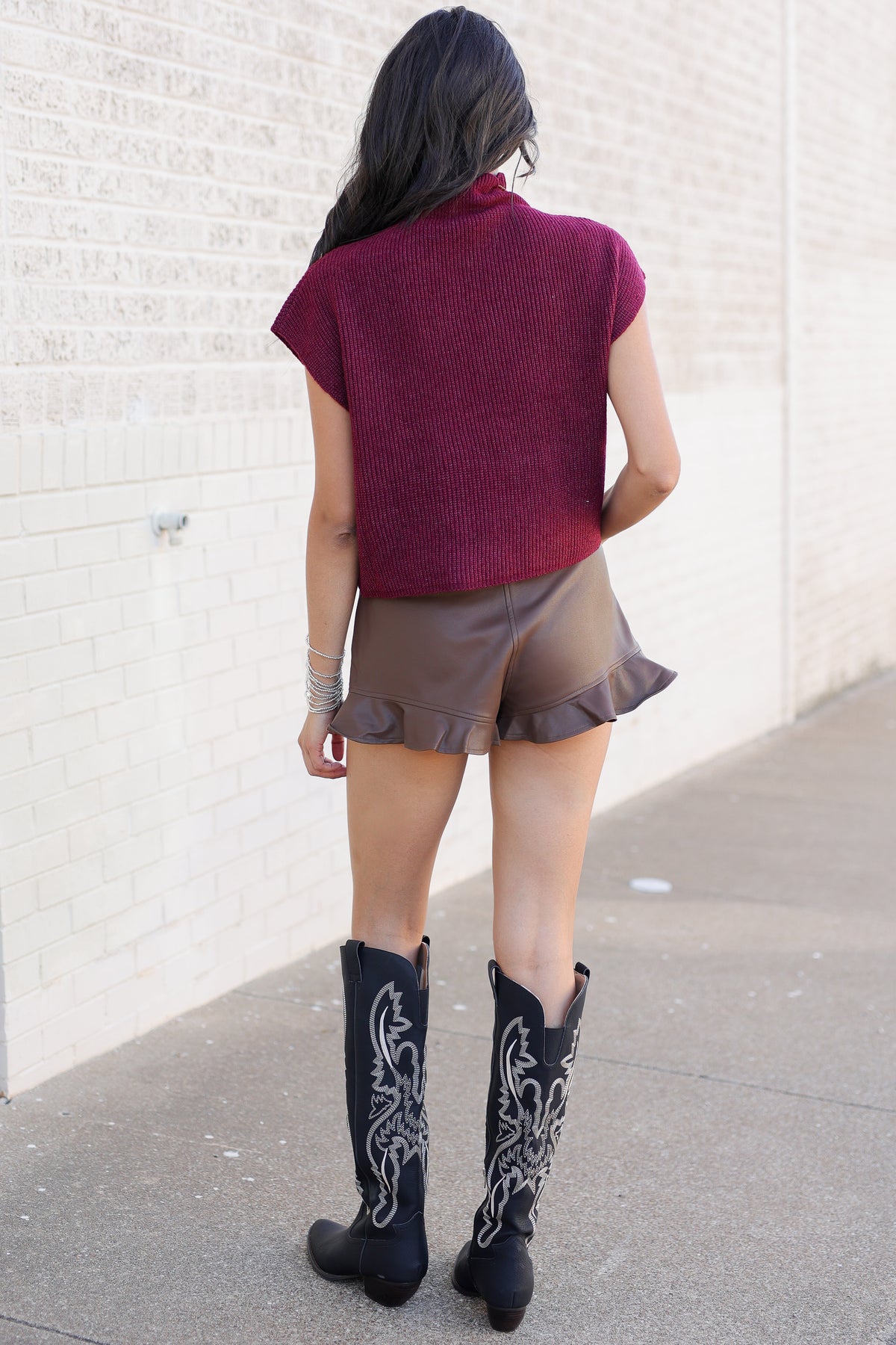 A woman photographed from behind wearing a textured burgundy short-sleeve mock neck top, brown ruffled shorts, and black western boots with white embroidery, standing against a light brick wall.