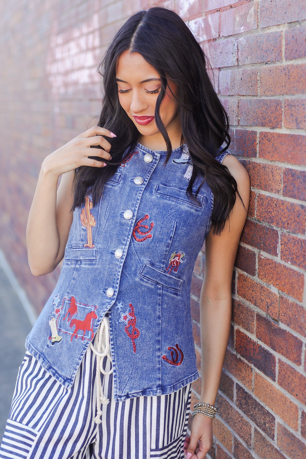 Woman wearing a denim vest with embroidery leaning against a brick wall.