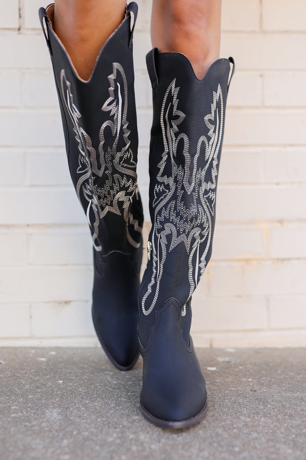 A pair of black leather cowboy boots with intricate silver embroidery on the shafts, worn by a model standing against a white brick wall.