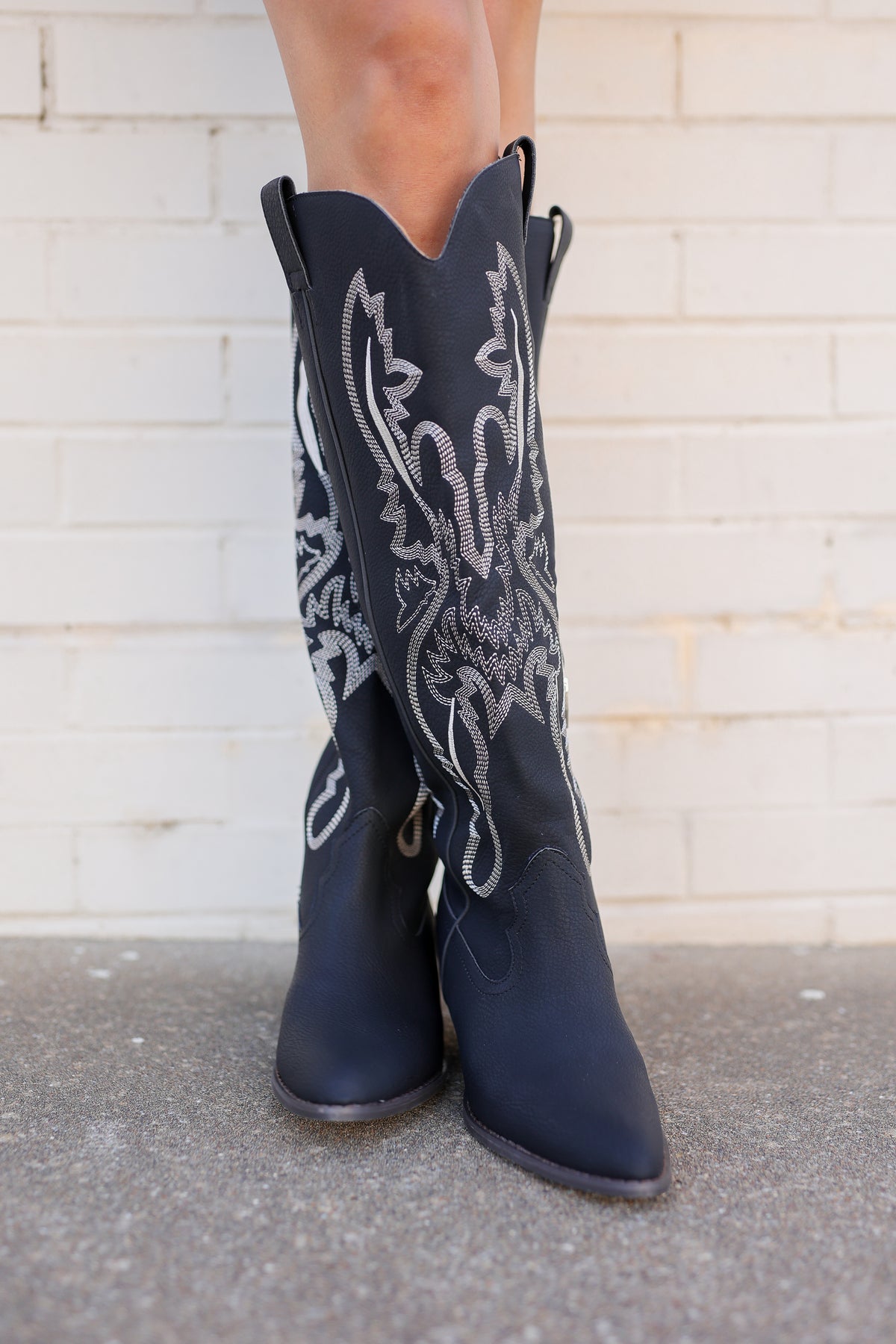 A pair of dark navy blue leather cowboy boots with white embroidered detailing on the shafts, shown on a model's feet against a white brick wall background.