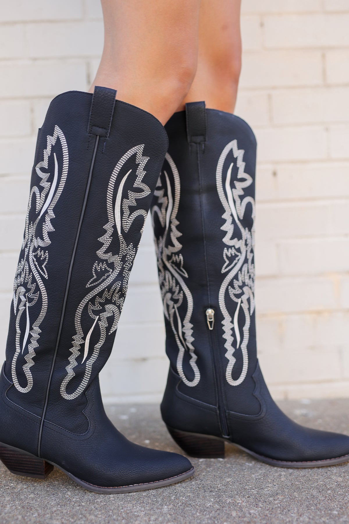 A pair of black leather cowboy boots with intricate white western embroidery, shown on a model's feet against a light brick wall.