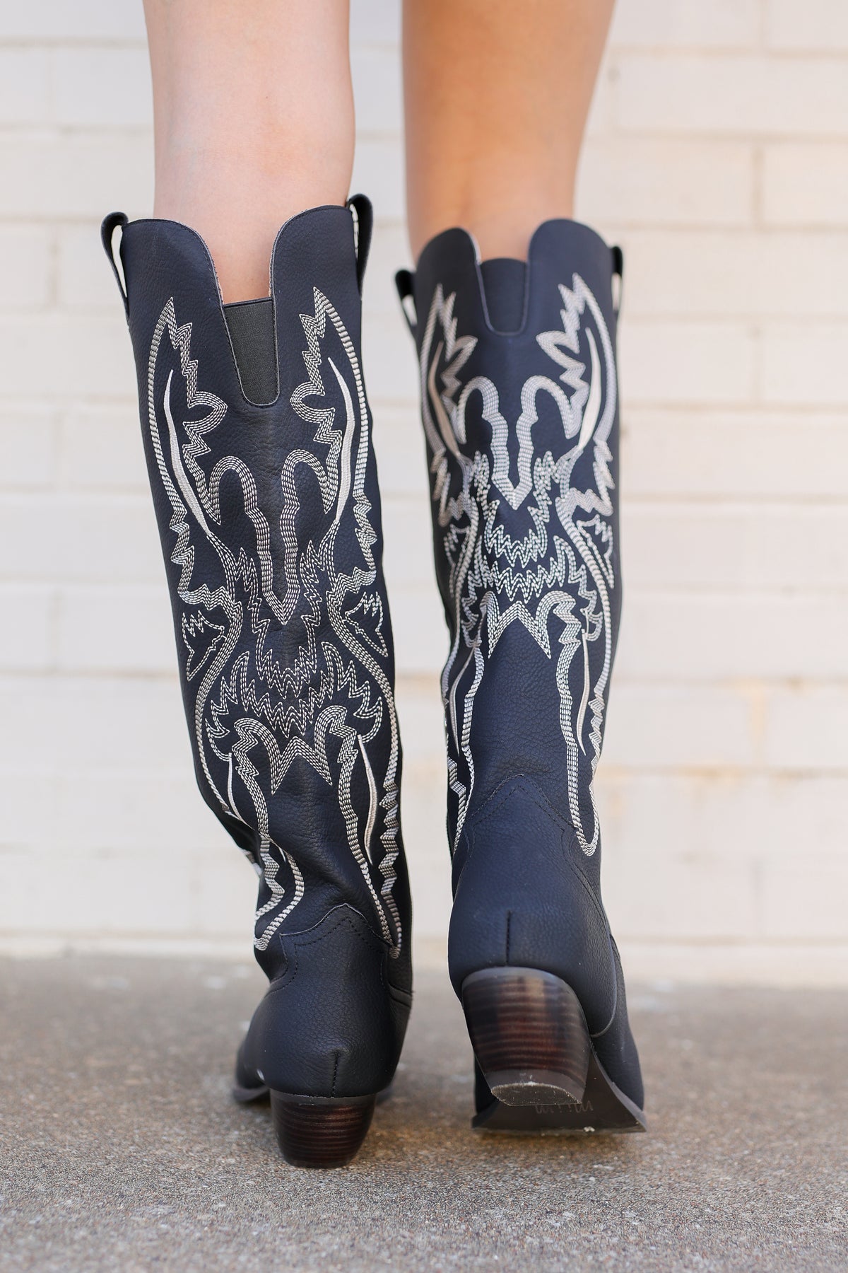 A pair of dark navy blue leather cowboy boots with intricate white western embroidery, shown from the back on a person's feet against a white brick wall.