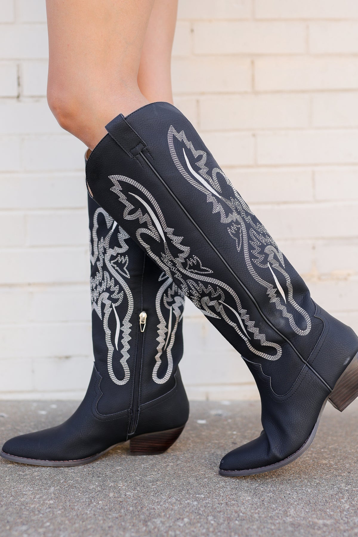 Black leather cowboy boots with intricate white and silver western embroidery, shown on a model's legs against a white brick wall background.