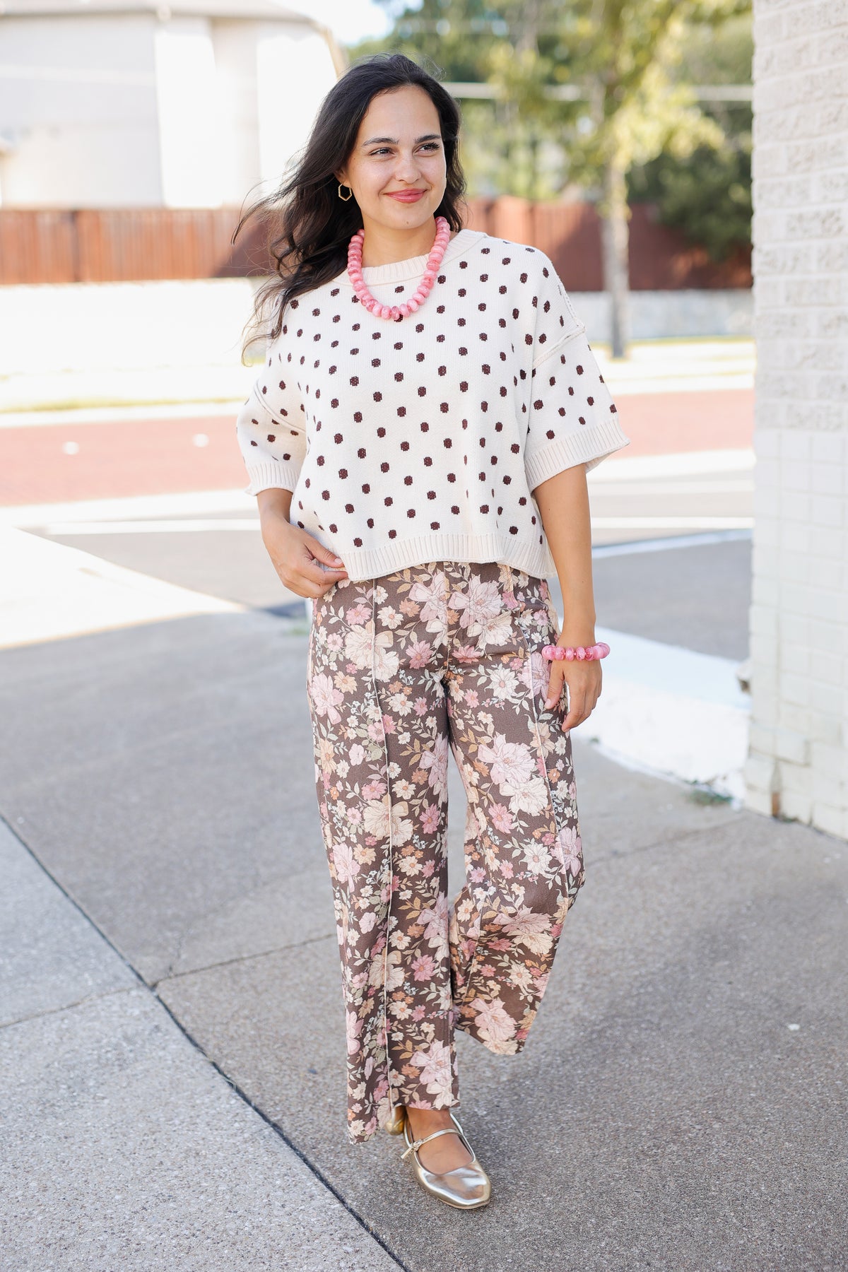 A woman posing outdoors on a sidewalk wearing a white short-sleeve top with black polka dots and matching wide-leg floral pants in pink, beige, and brown tones.