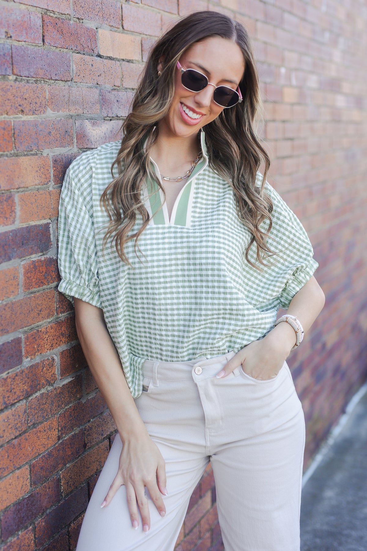 Woman wearing a green checkered shirt and beige pants against a brick wall.