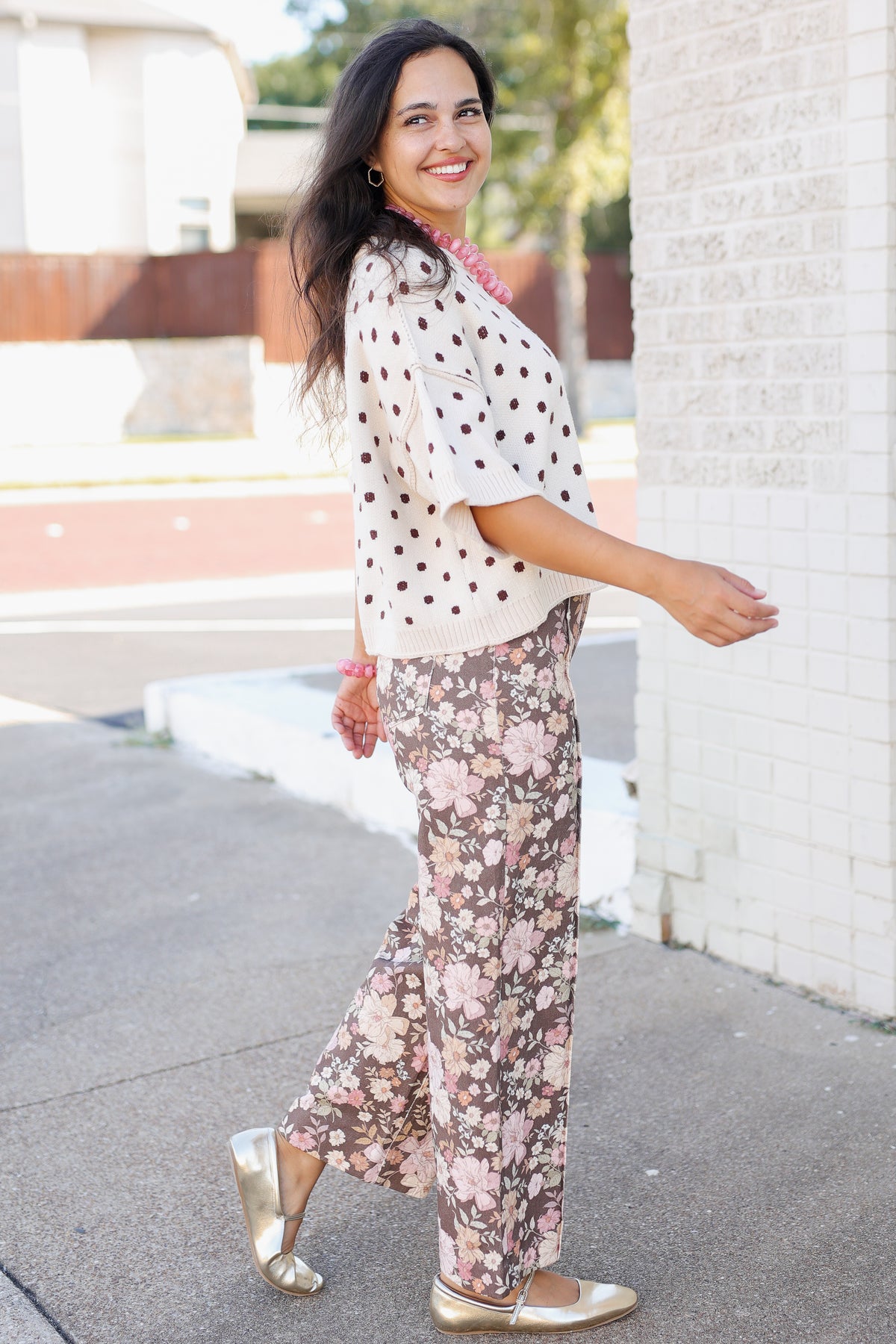A woman posing outdoors on a sidewalk next to a white brick wall, wearing a cream short-sleeve top with black polka dots and matching floral wide-leg pants in pink and beige tones, paired with metallic gold flats.