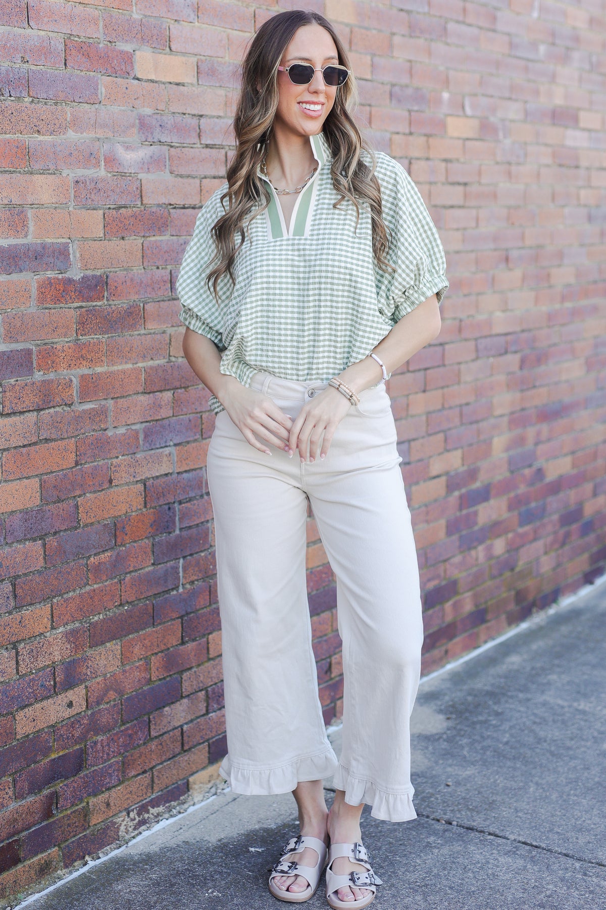 Woman wearing a green checkered shirt and white pants standing against a brick wall.