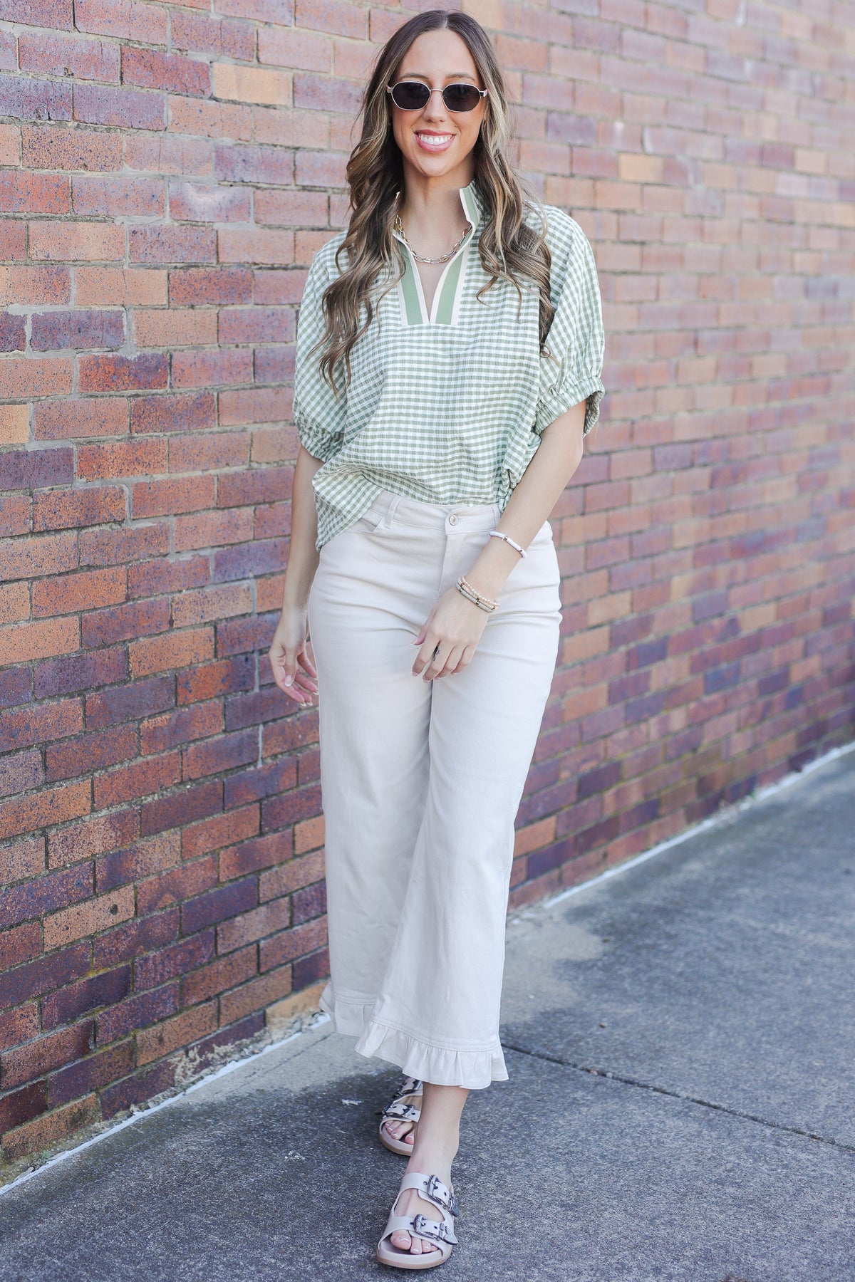Woman wearing a green checkered shirt and white pants against a brick wall.