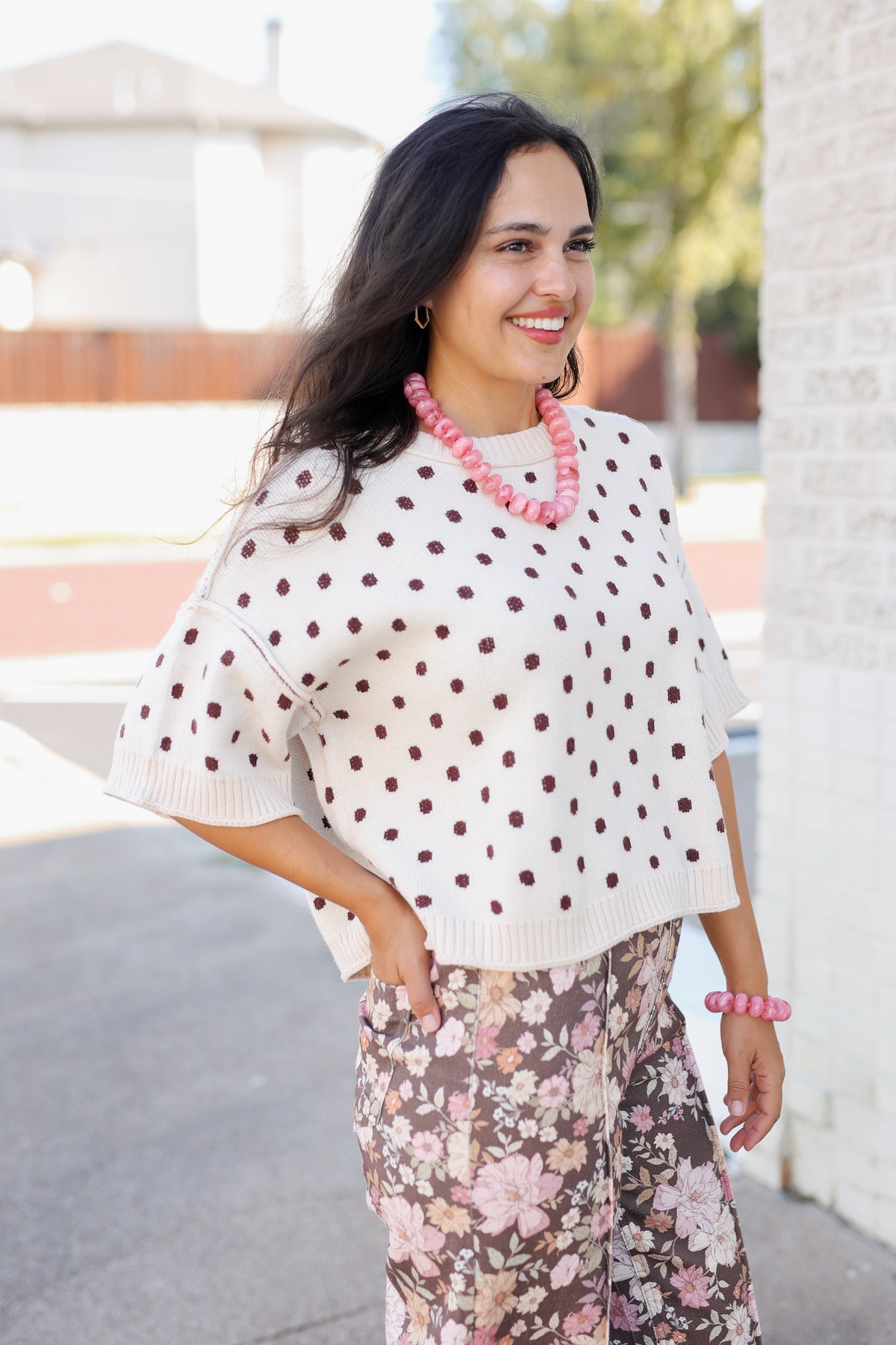 A woman wearing a cream knit sweater with dark brown polka dots and floral print pants, accessorized with a chunky pink beaded necklace and bracelet, standing outdoors.