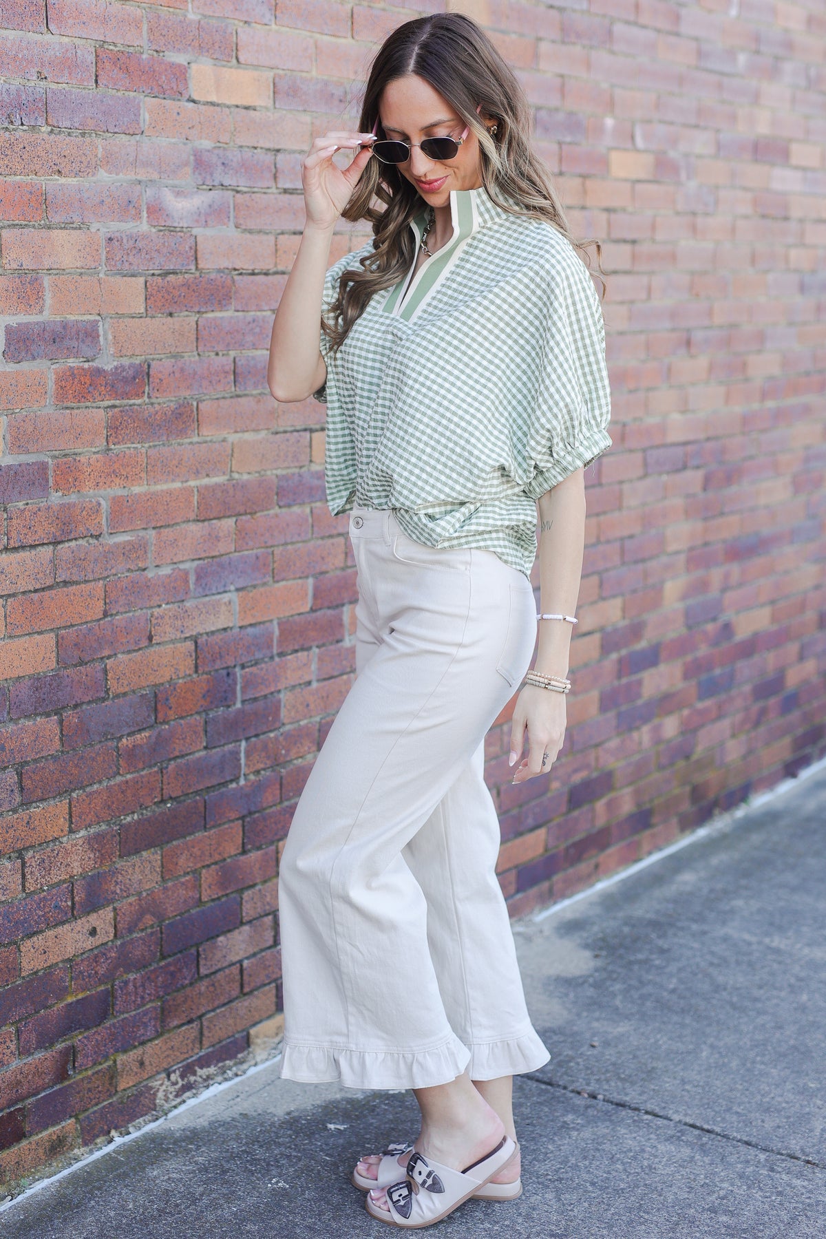 Woman wearing a green patterned blouse and white pants against a brick wall.