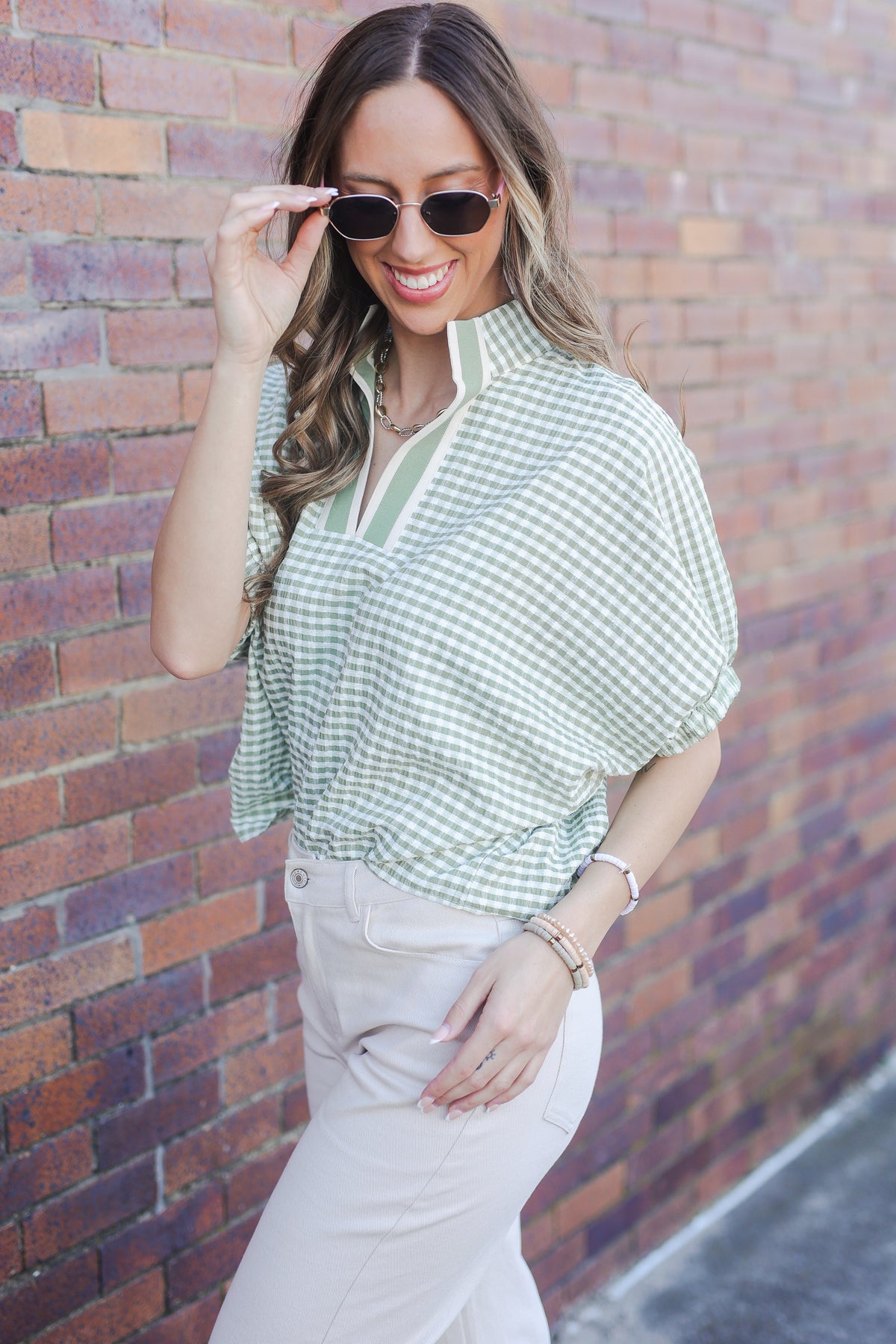 Woman wearing a green checkered top and white pants against a brick wall.