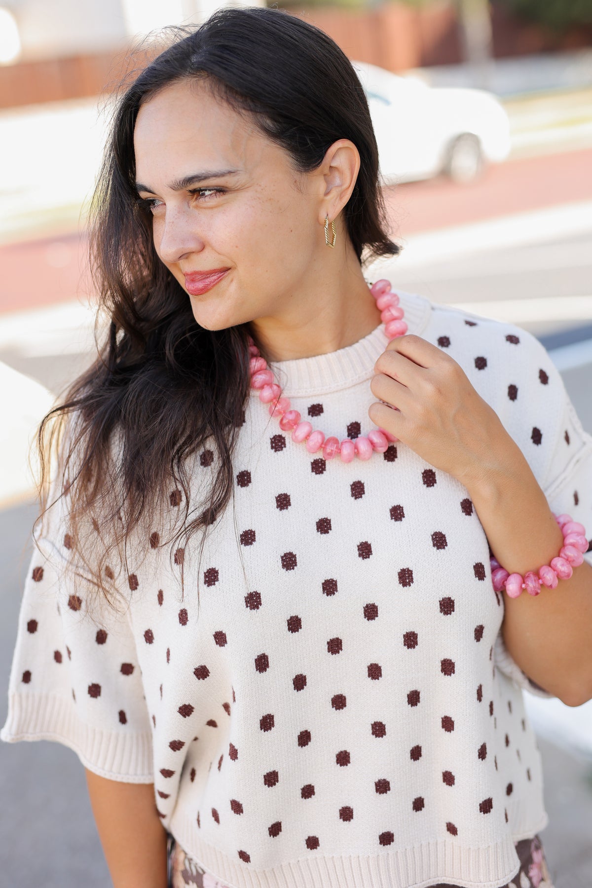 A woman wearing a cream knit sweater with dark brown polka dots, styled with a chunky pink beaded necklace and bracelet, photographed outdoors on a blurred street background.