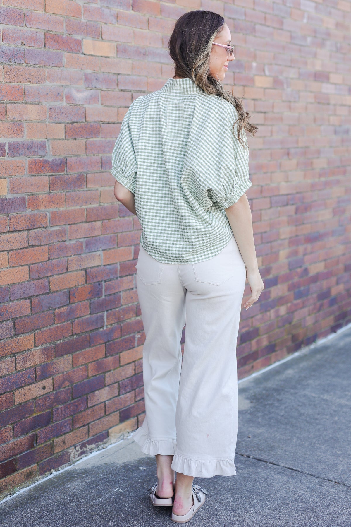 Woman wearing a green checkered shirt and beige pants against a brick wall.