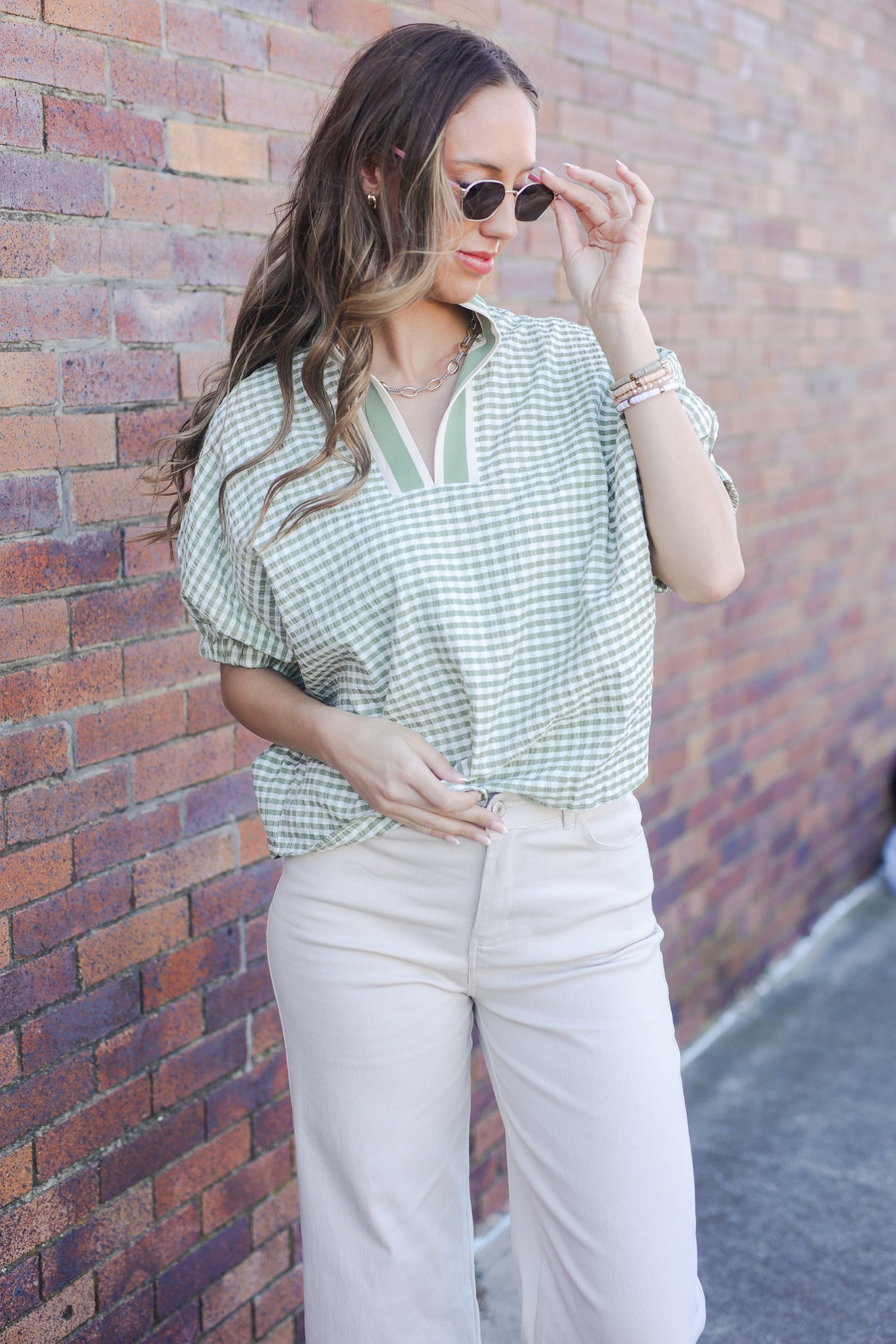 Woman wearing a green checkered shirt and white pants against a brick wall.