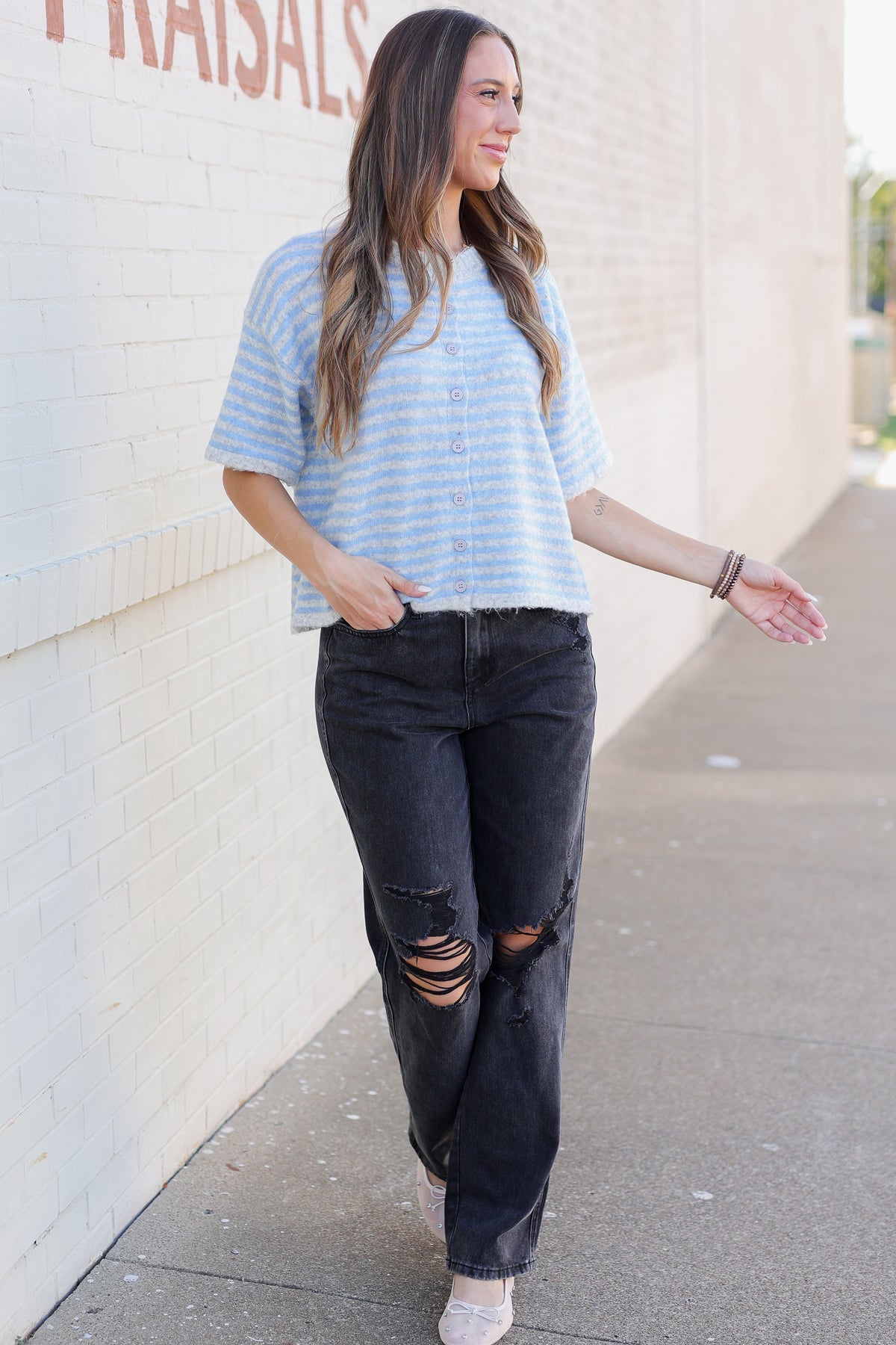 A woman walking outdoors wearing a light blue and white striped short-sleeve t-shirt, black distressed jeans, and white sneakers, standing next to a white brick wall.