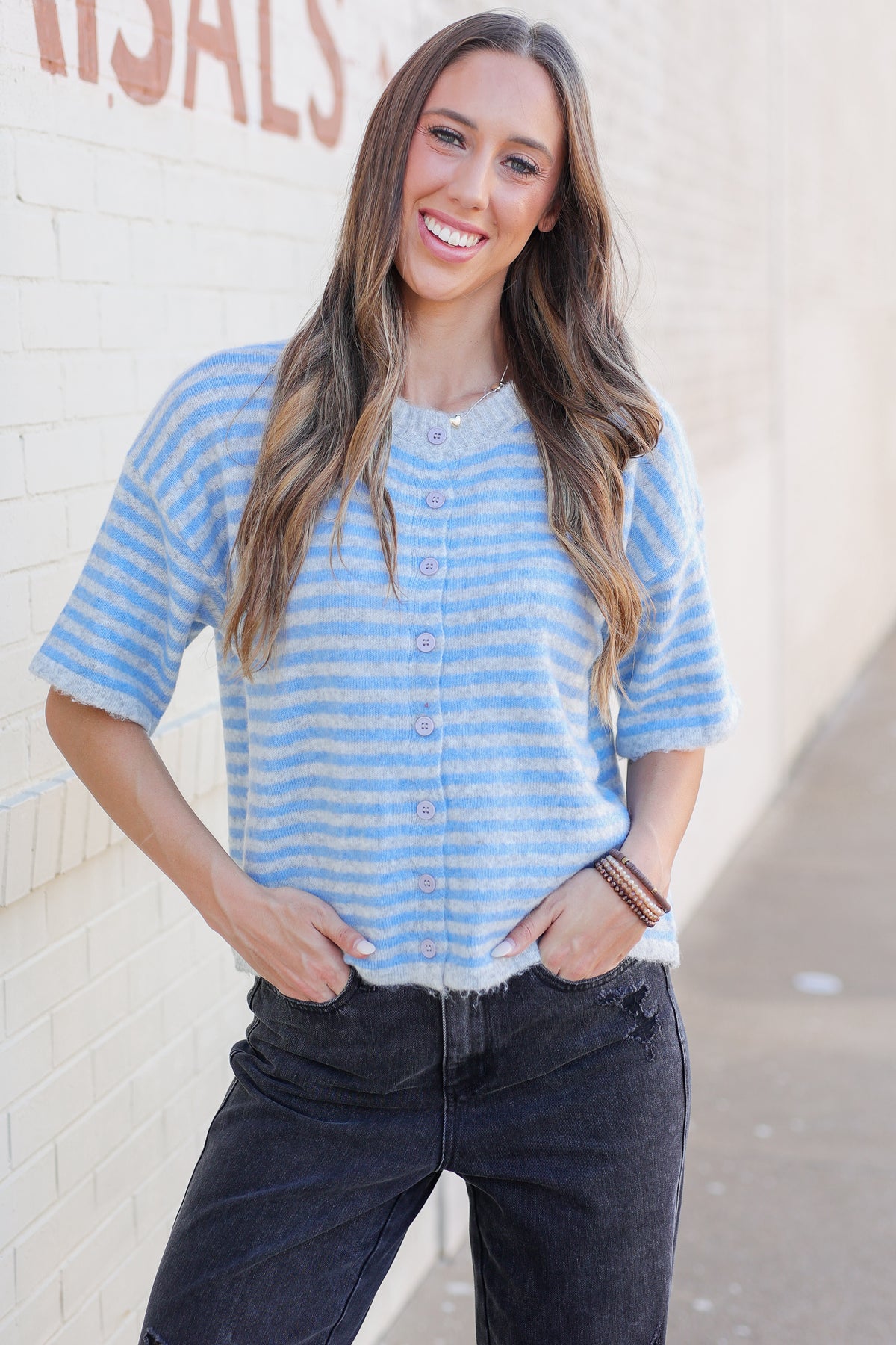 A woman smiling and posing outdoors wearing a light blue and white striped short-sleeve button-up cardigan with dark gray jeans.