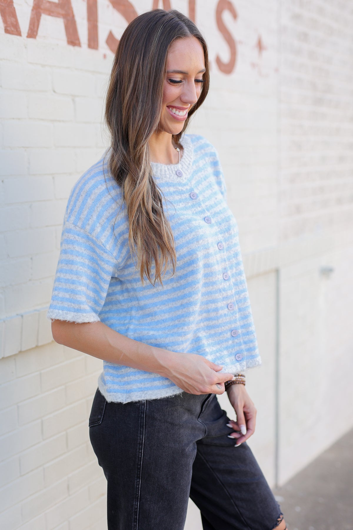 A woman wearing a light blue and white striped short-sleeve knit top with buttons, paired with dark jeans, standing against a white brick wall.