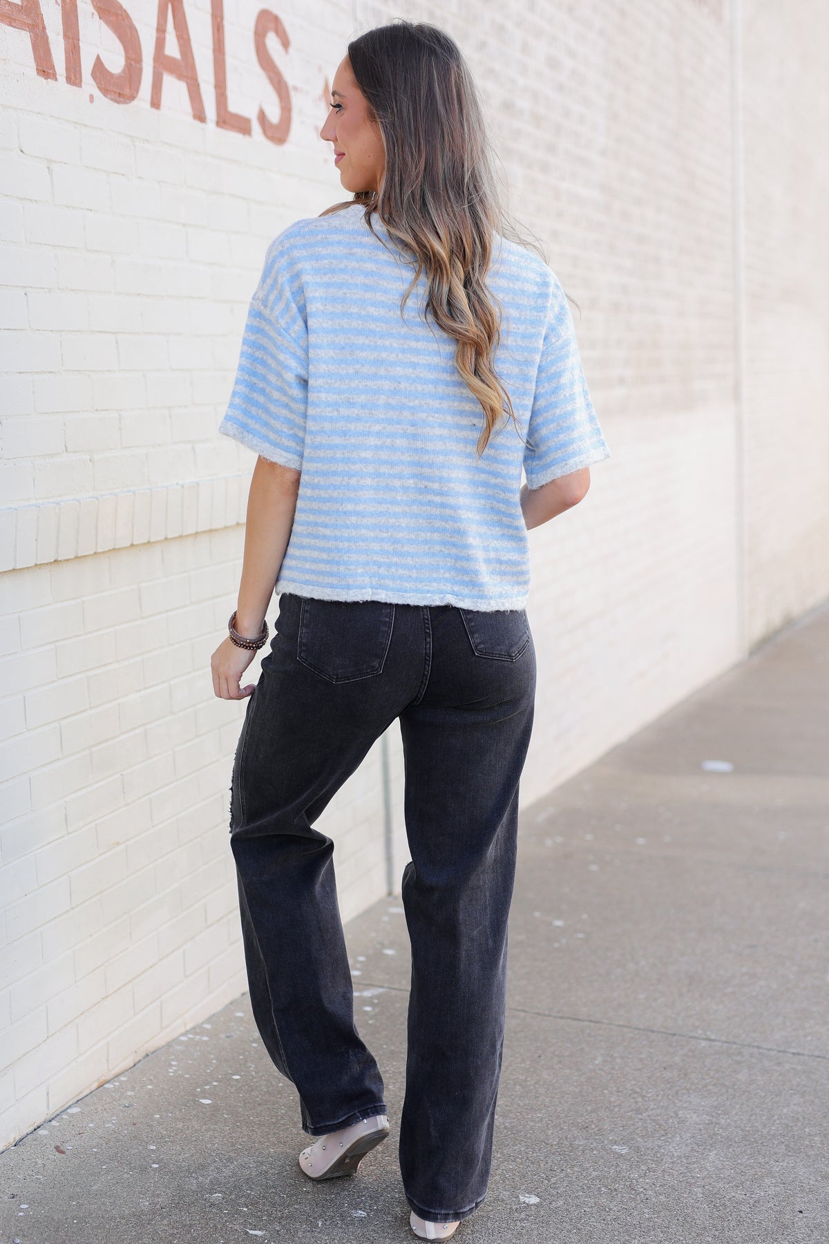 A woman is shown from behind wearing a light blue and white striped short-sleeve t-shirt, dark wash jeans, and white sneakers, standing on a sidewalk next to a white brick wall.