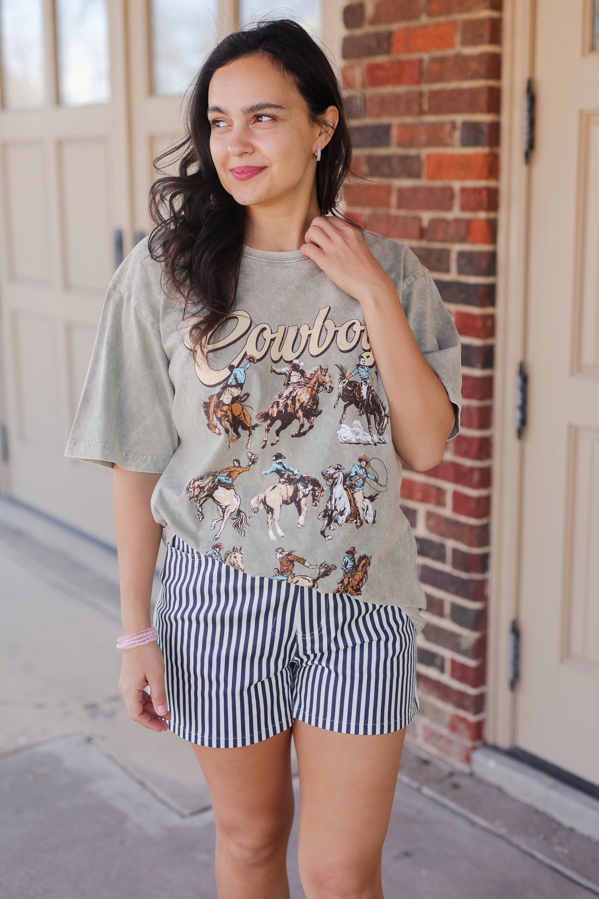 Woman wearing a graphic t-shirt with 'Cowboy' and horse illustrations, standing in front of a brick wall.