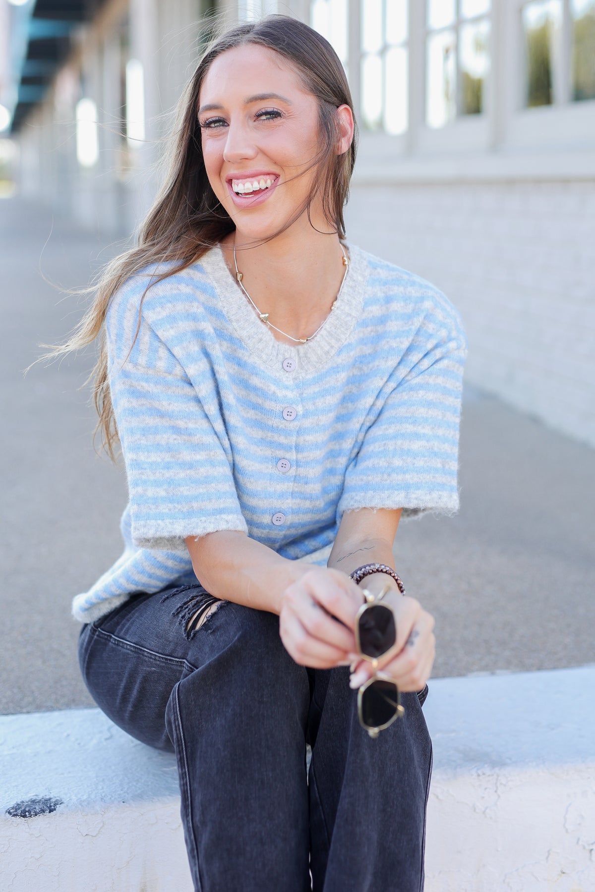 A woman sitting outdoors on a concrete ledge wearing a light blue and white striped short-sleeve knit top with buttons, paired with dark jeans.